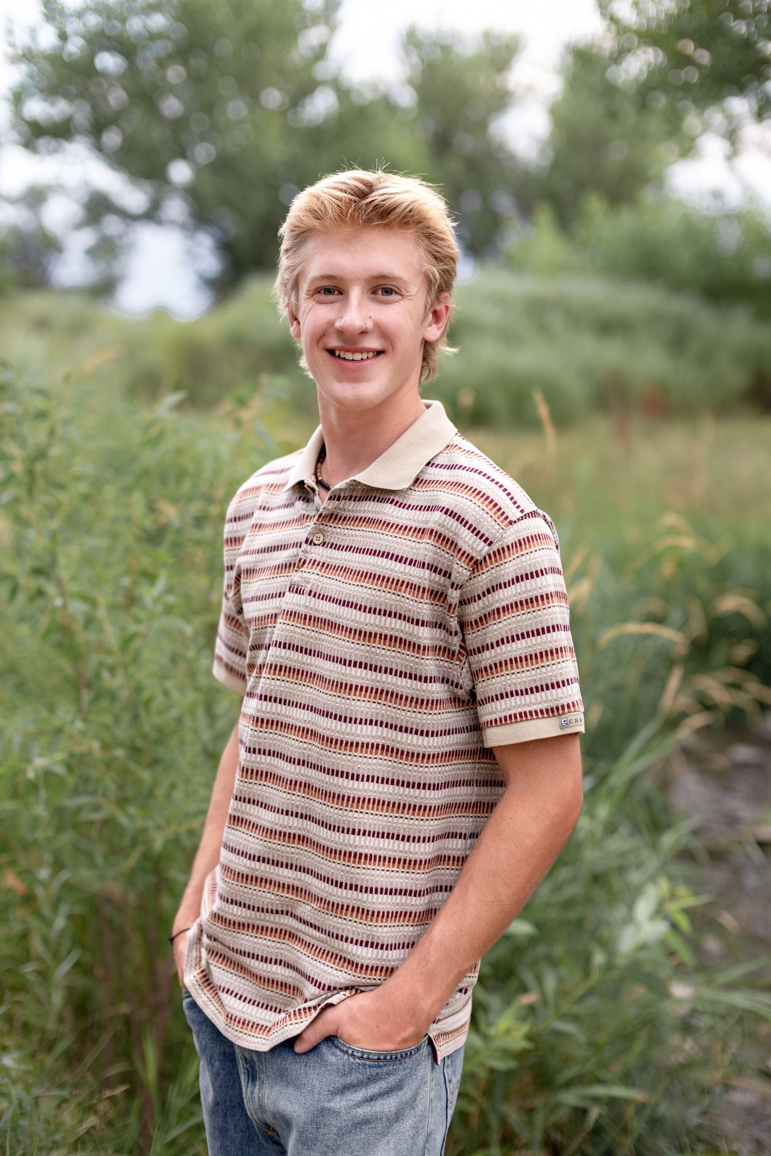 Smiling teenage boy with blonde hair wearing a striped polo shirt, standing outdoors among tall green plants.