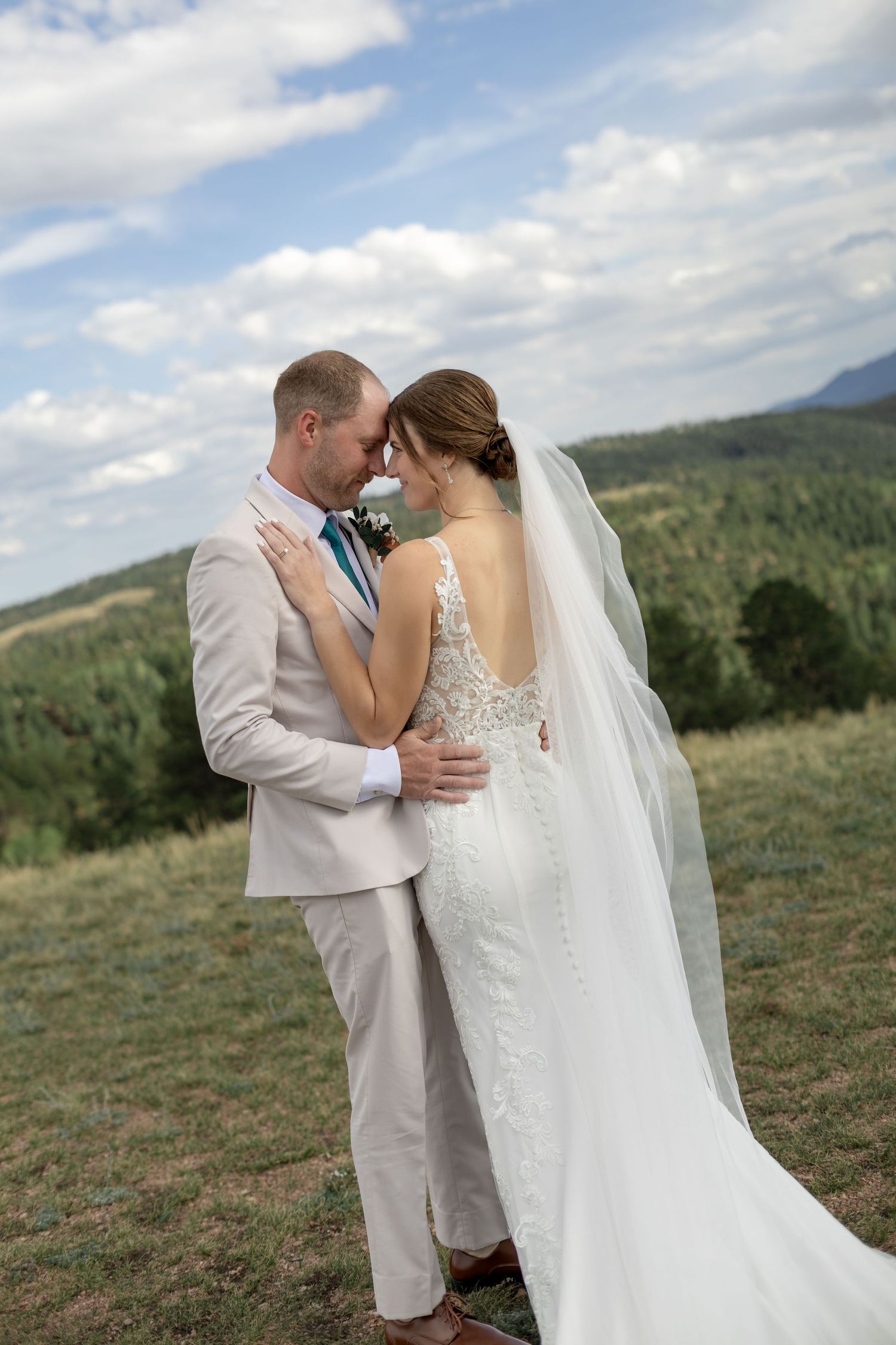 Bride and groom embrace in a meadow with mountain views, she in lace gown with veil, he in beige suit.