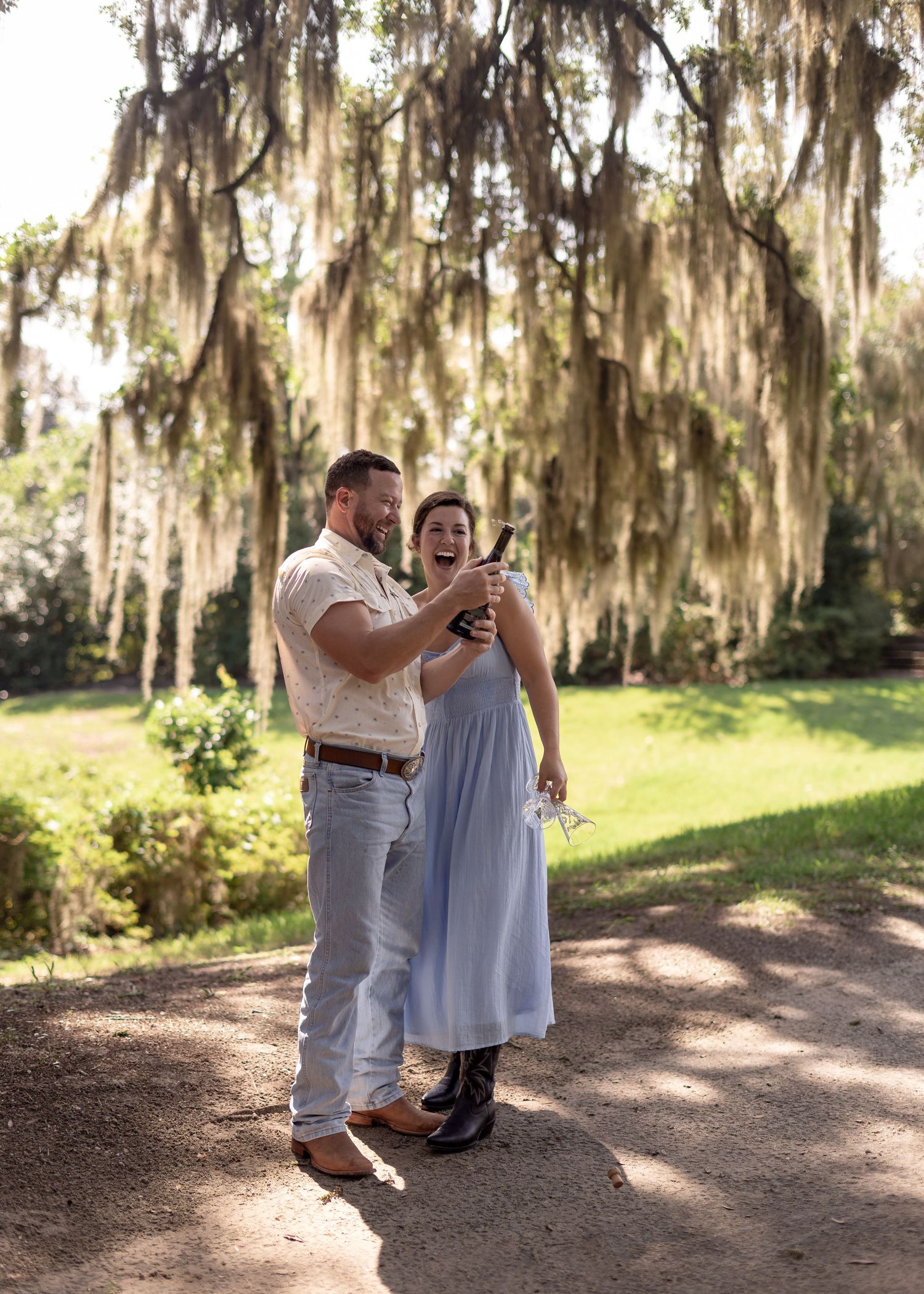 A happy couple celebrates outdoors under Spanish moss trees, sharing champagne in a sunny park setting.