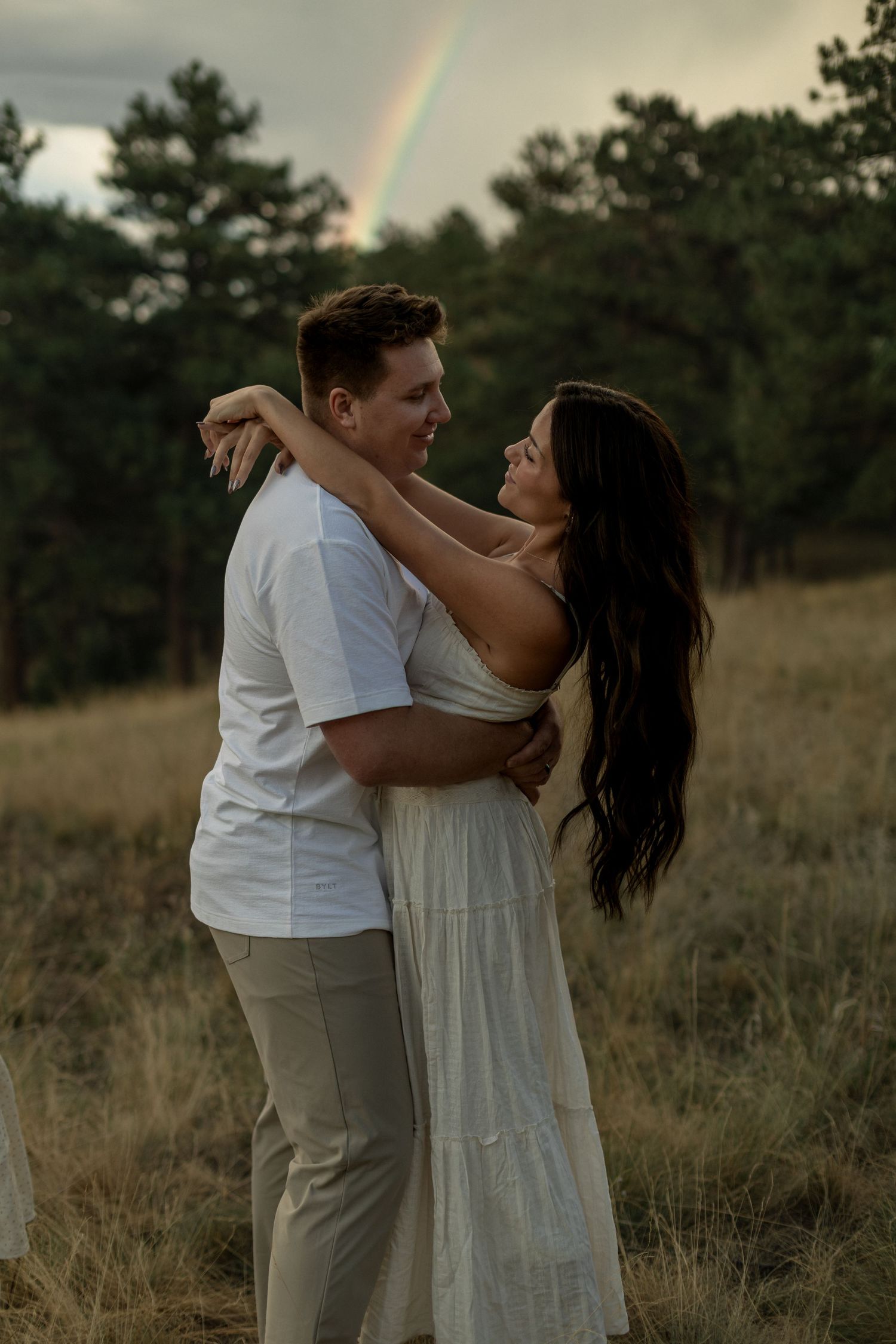 Couple embracing in a golden field with a rainbow and pine trees in the background at dusk.