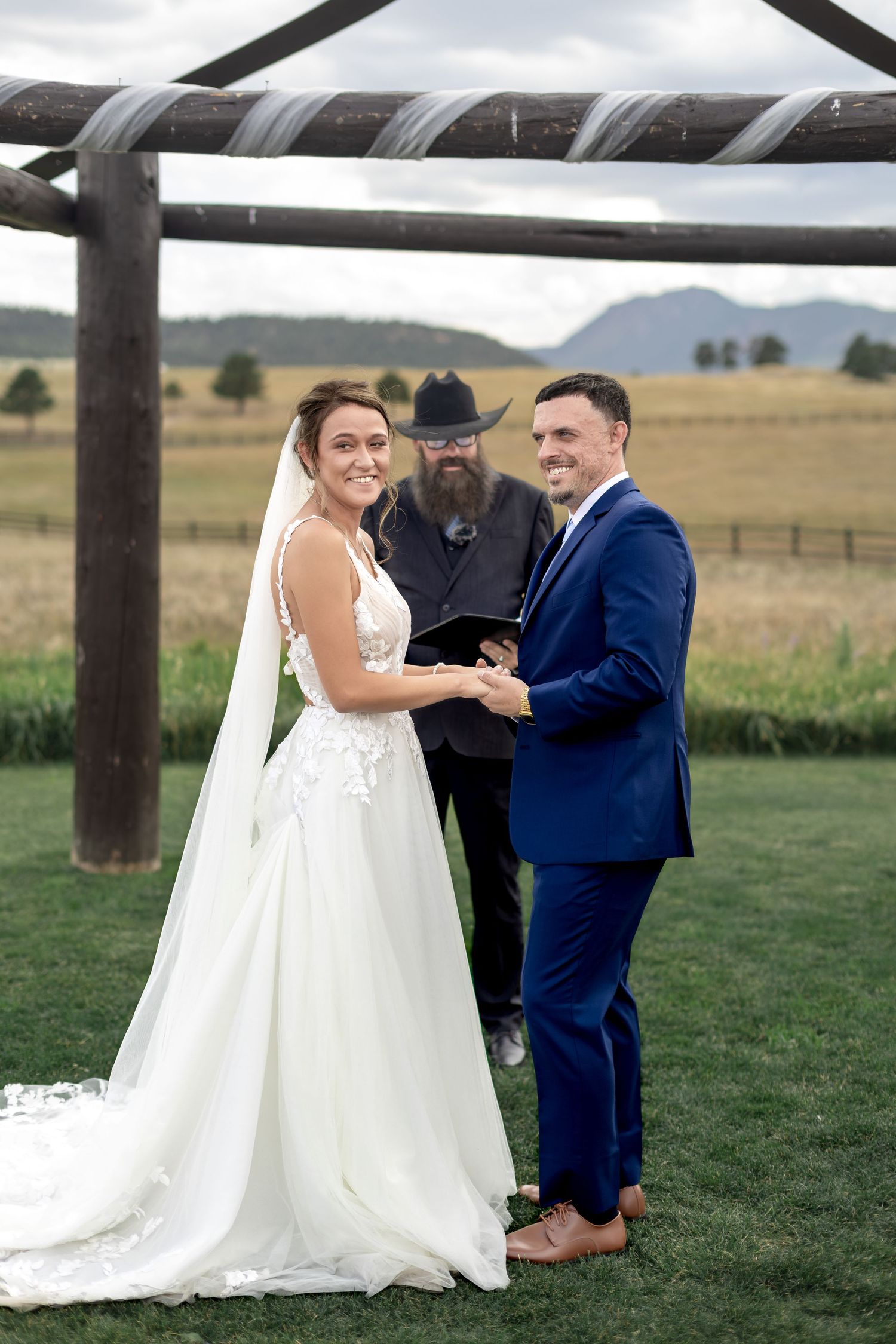 Smiling bride and groom holding hands during outdoor wedding ceremony with mountain backdrop.