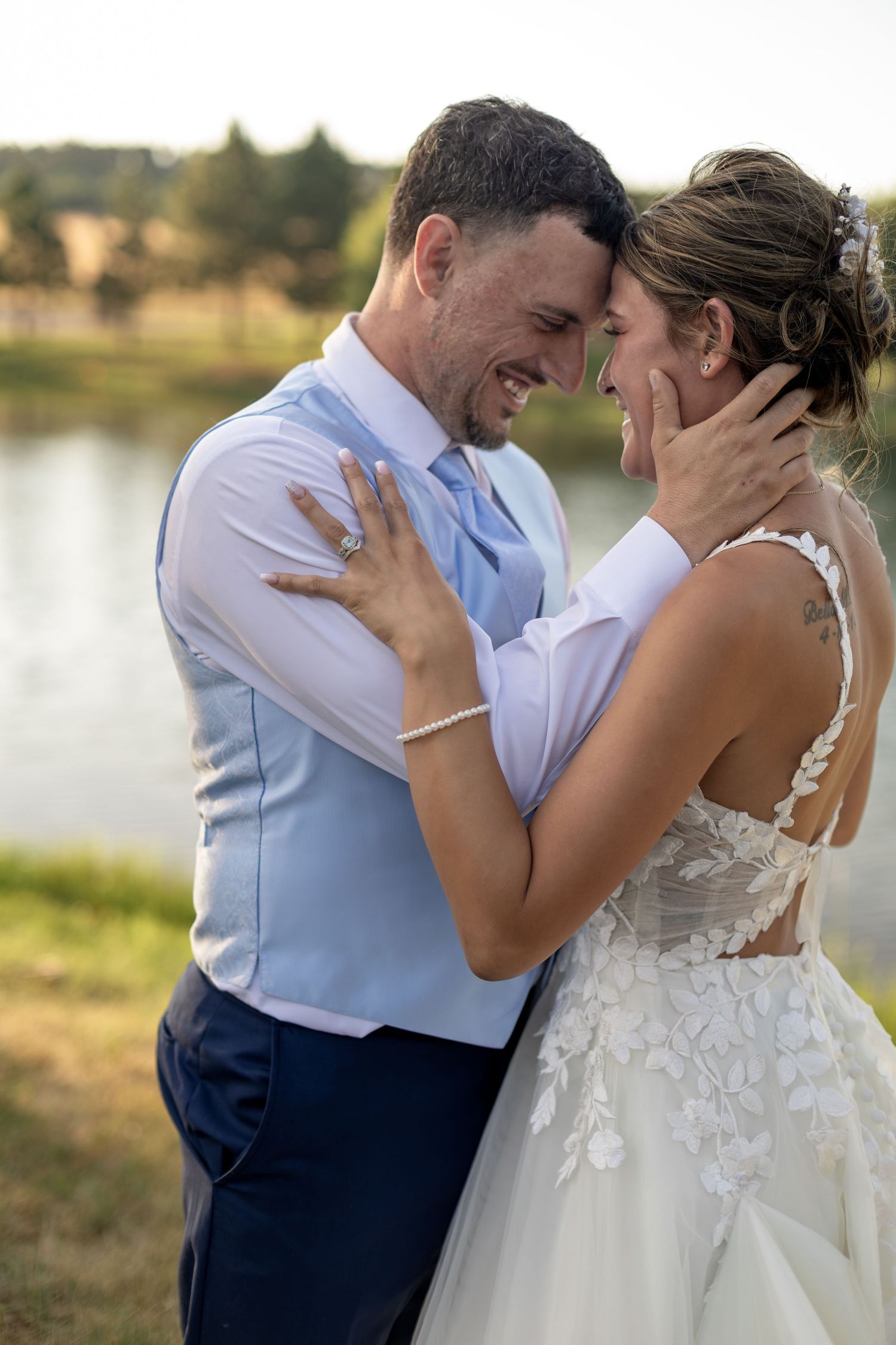 Laughing bride and groom holding each other tenderly near a sunlit lake outdoors.
