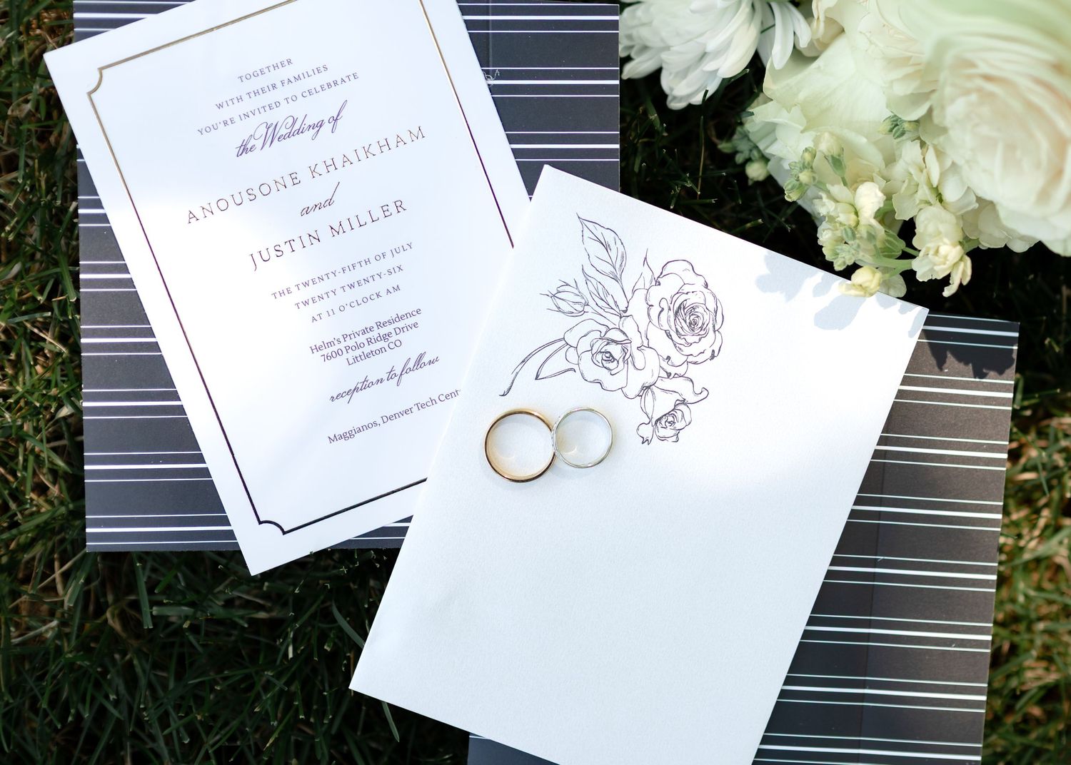 Elegant wedding invitation suite with gold rings and floral details on striped background with white flowers.