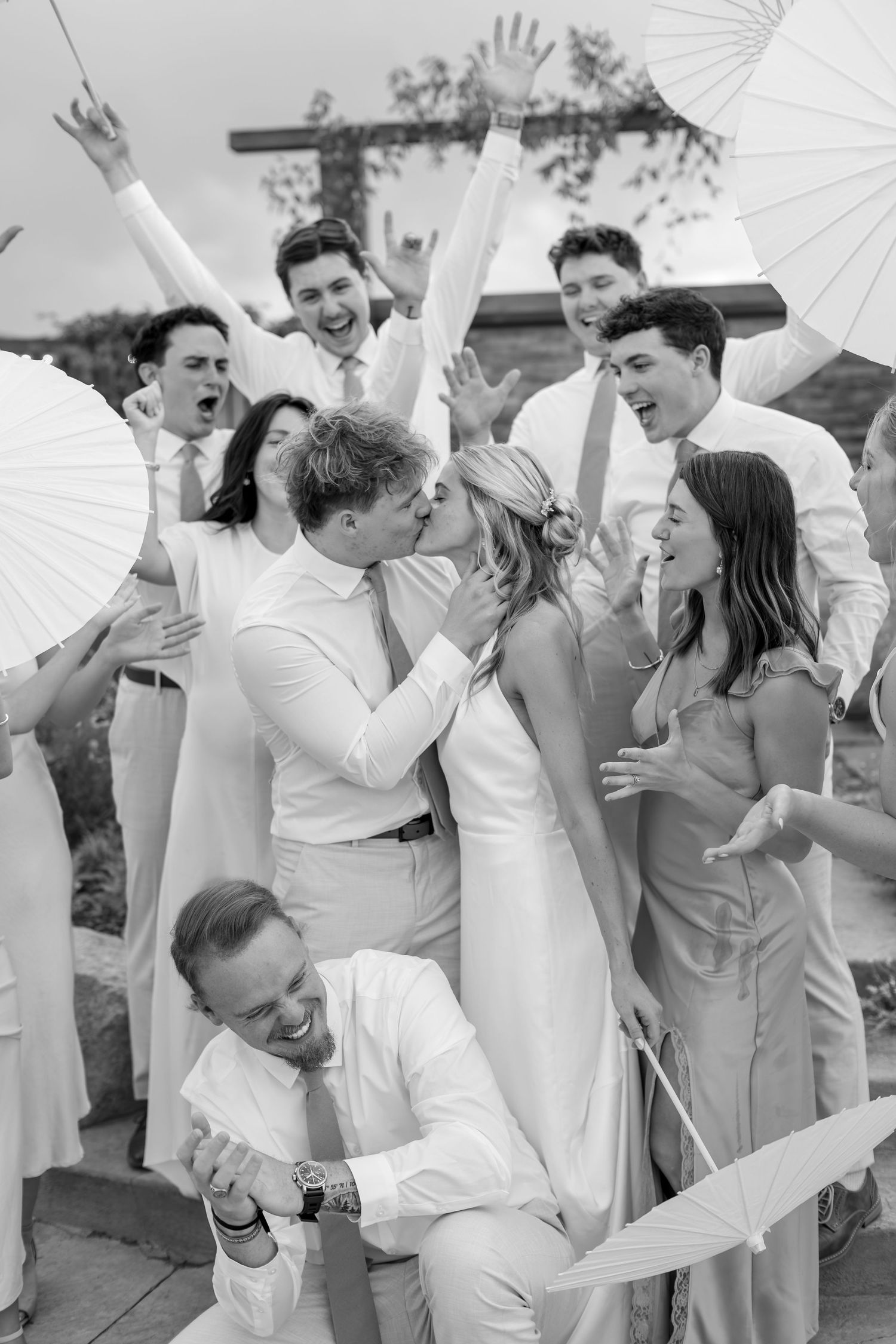 Black and white photo of bride and groom kissing surrounded by cheering wedding party holding parasols.
