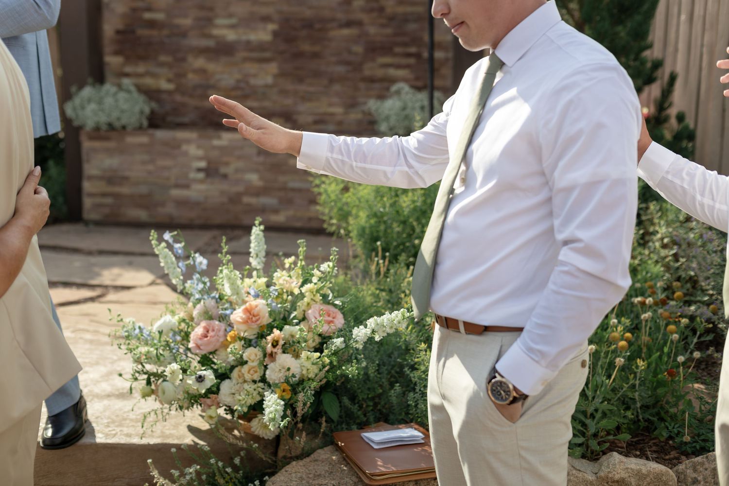 Man in white shirt and sage tie gestures near elegant floral wedding bouquet with peach and white blooms outdoors.