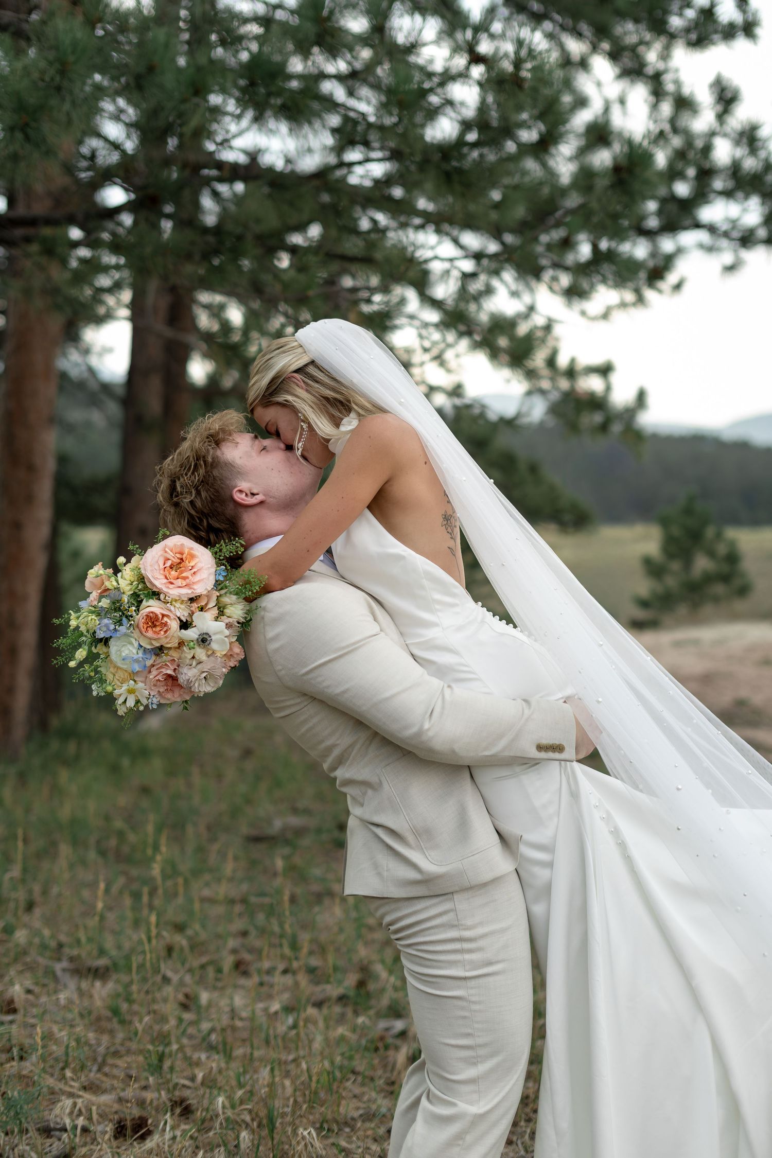 Groom in ivory suit lifts bride in white gown and veil for a romantic kiss outdoors near pine trees.