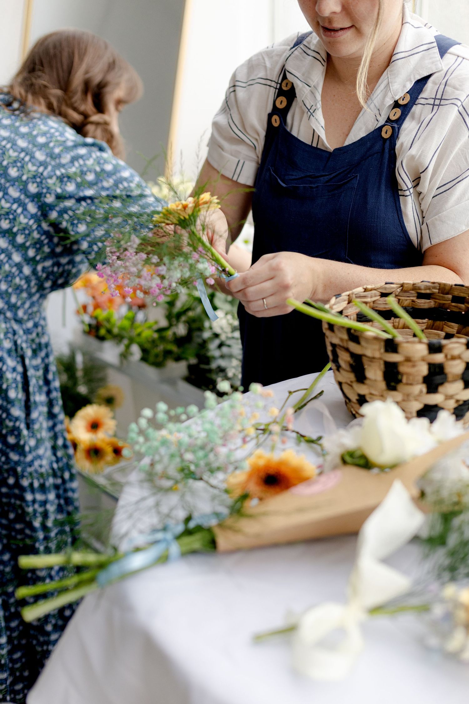 Florist in apron arranging fresh wildflowers at a white table with a wicker basket and colorful bouquets.