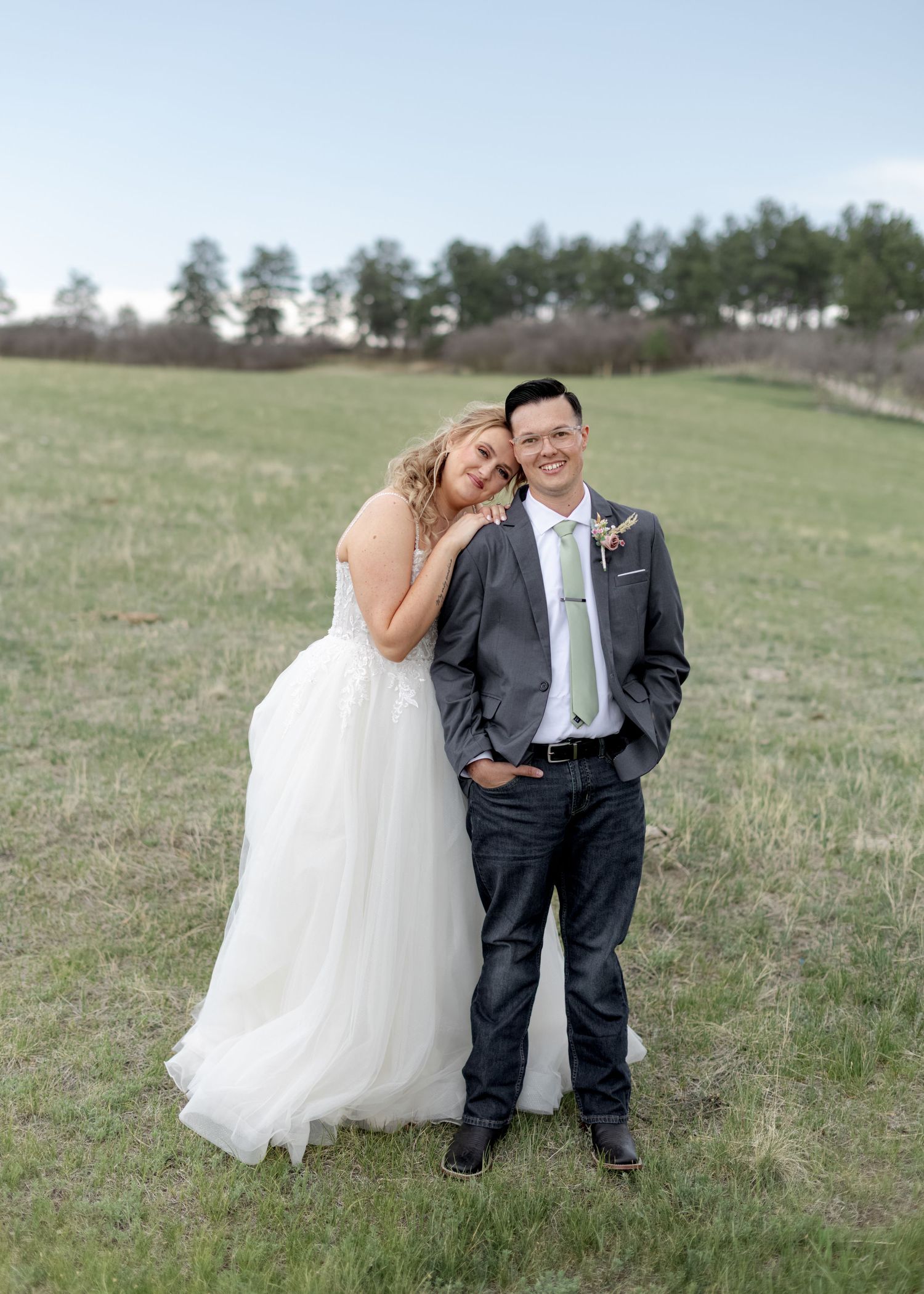 Smiling bride in white tulle gown leans on groom in gray suit standing in green meadow.