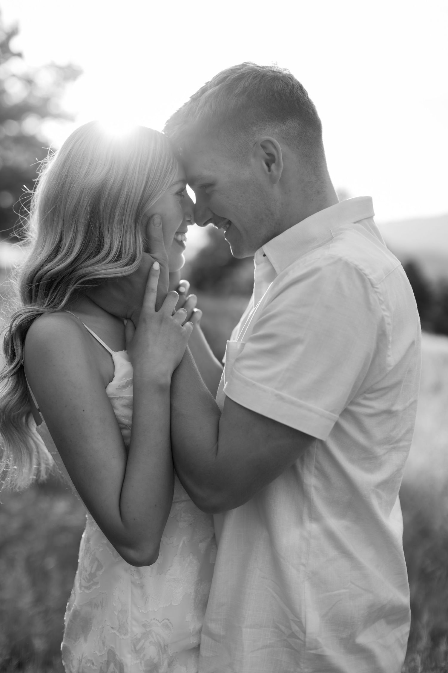 Black and white photo of a romantic couple touching foreheads and holding hands outdoors in a sunlit field.