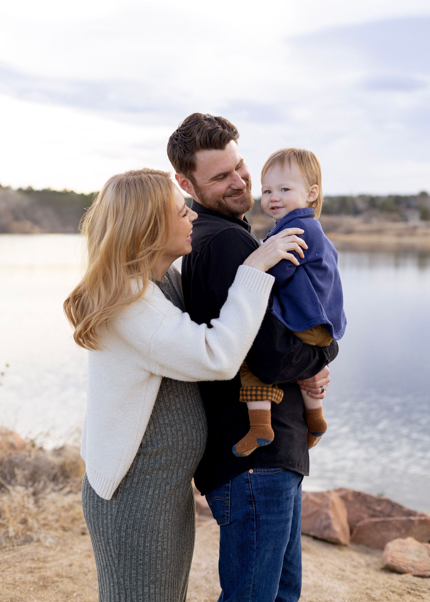 A happy young family with a toddler poses by a lakeside, with the parents smiling and holding their child outdoors.