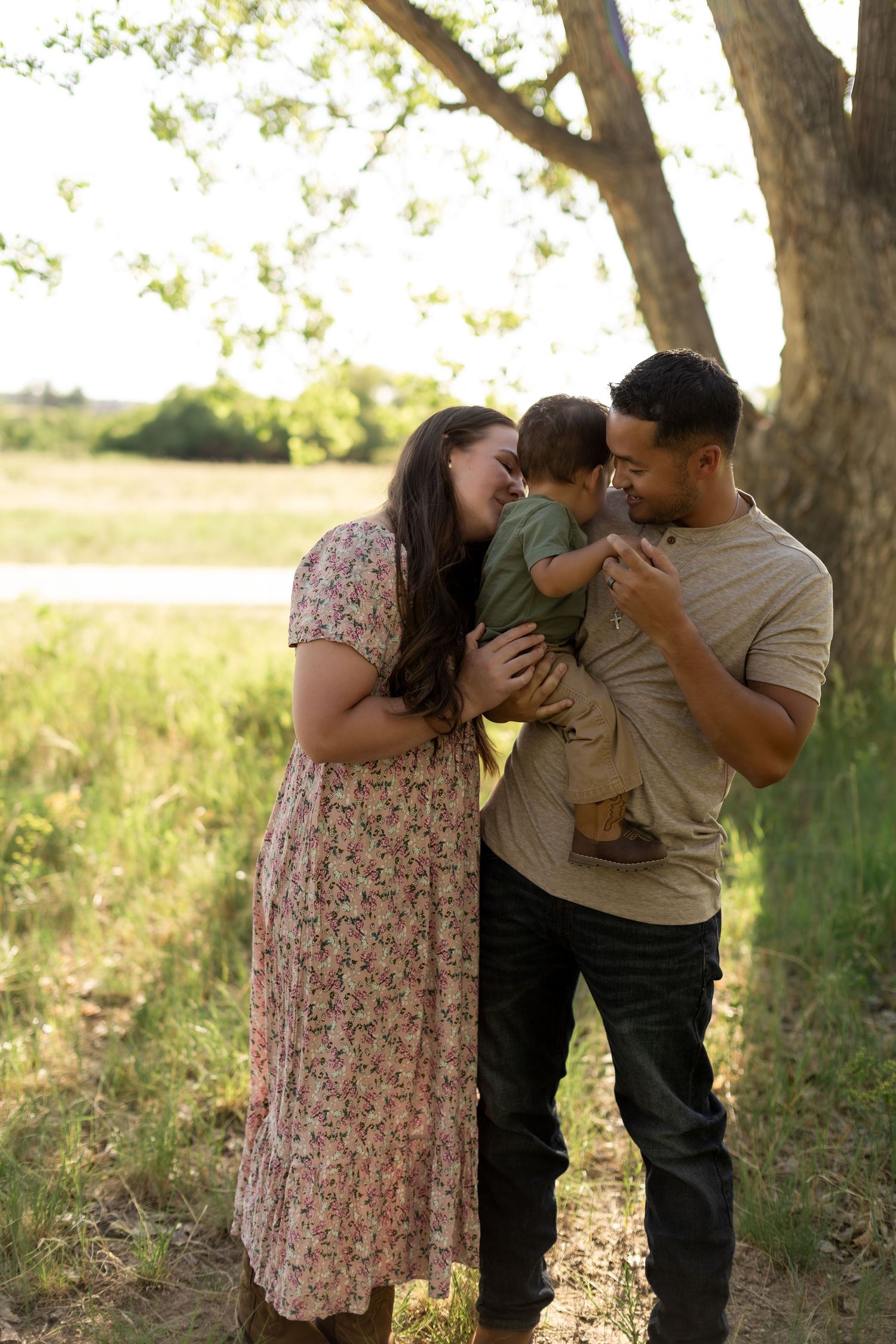 A happy family of three shares a tender moment outdoors, with parents smiling at their toddler near a tree.