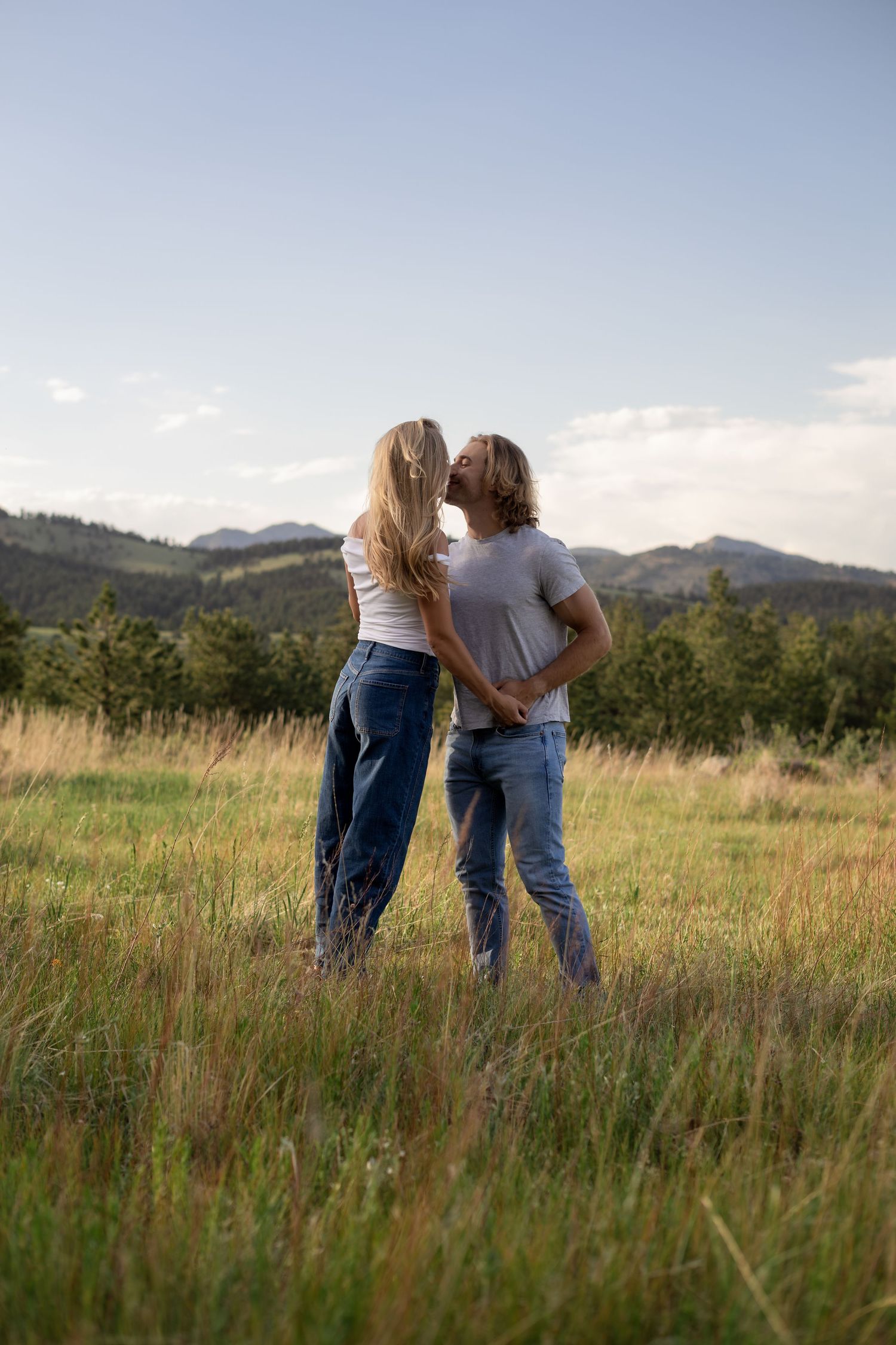 Couple sharing a kiss in a scenic mountain meadow, both wearing white tops and jeans.