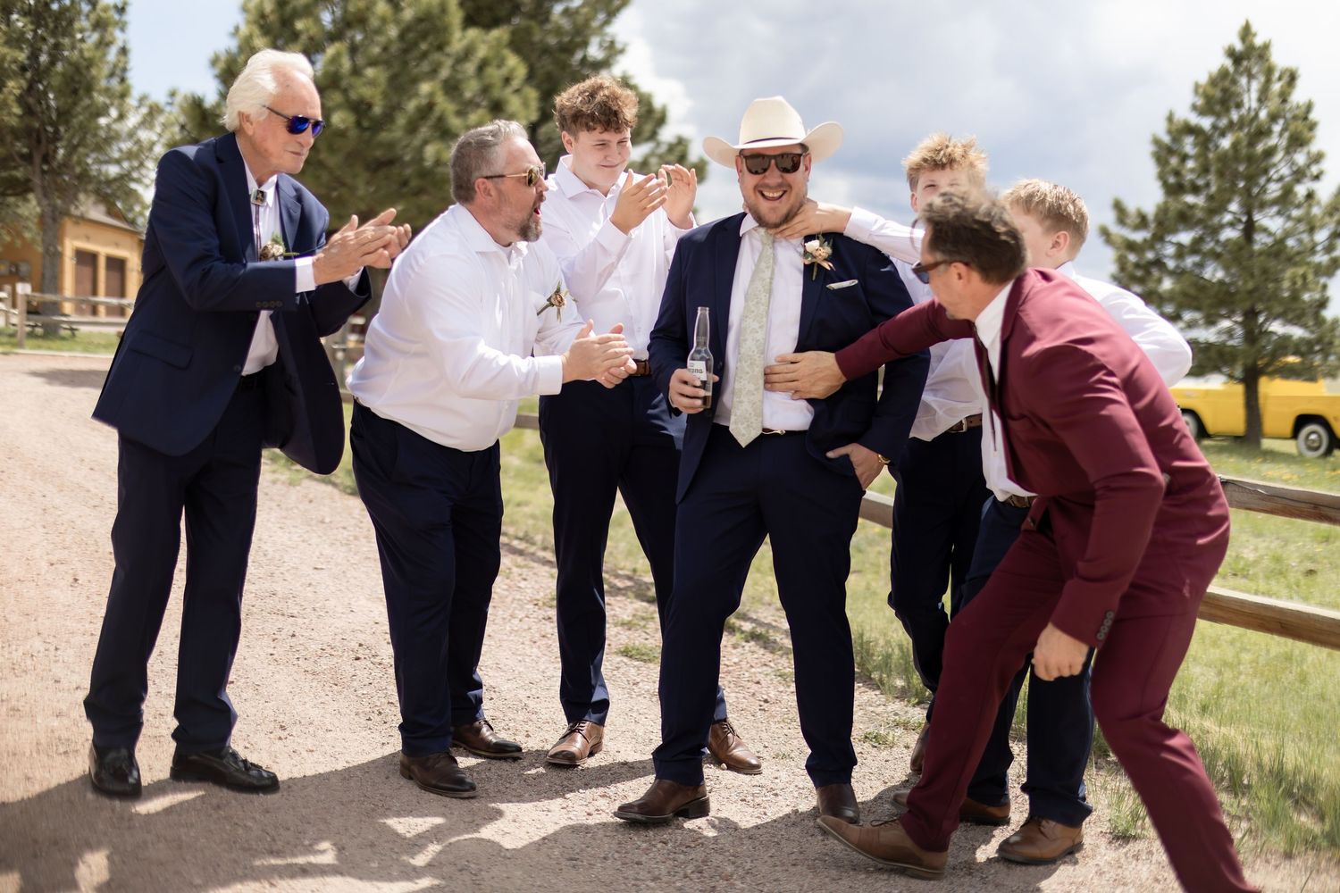 Groomsmen laughing and celebrating outdoors at a wedding, wearing navy suits and burgundy tuxedo.