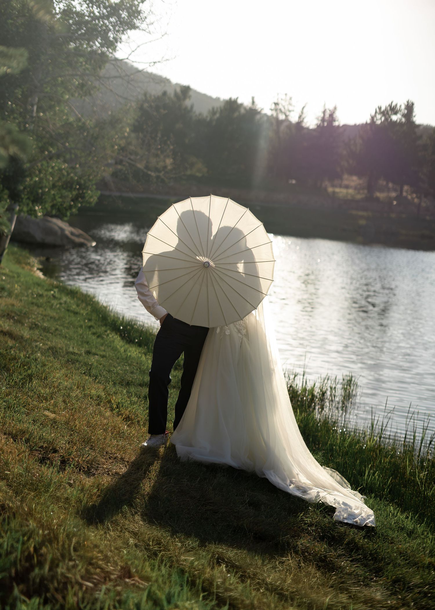 Wedding couple holding a white parasol umbrella by a serene lake, bride in flowing gown, lush greenery backdrop.