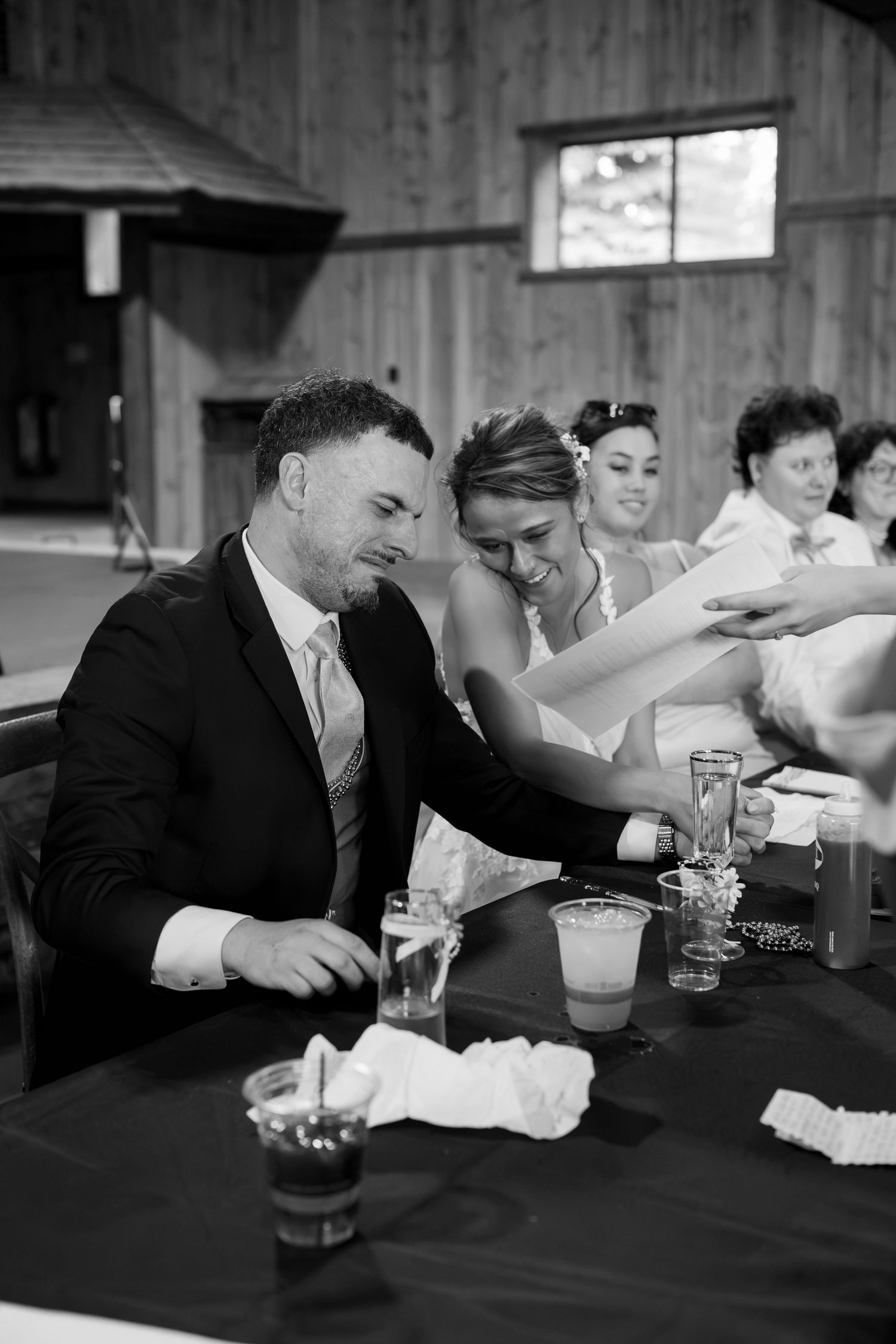 Black and white photo of newlyweds reviewing papers at their wedding reception table inside a rustic barn venue.