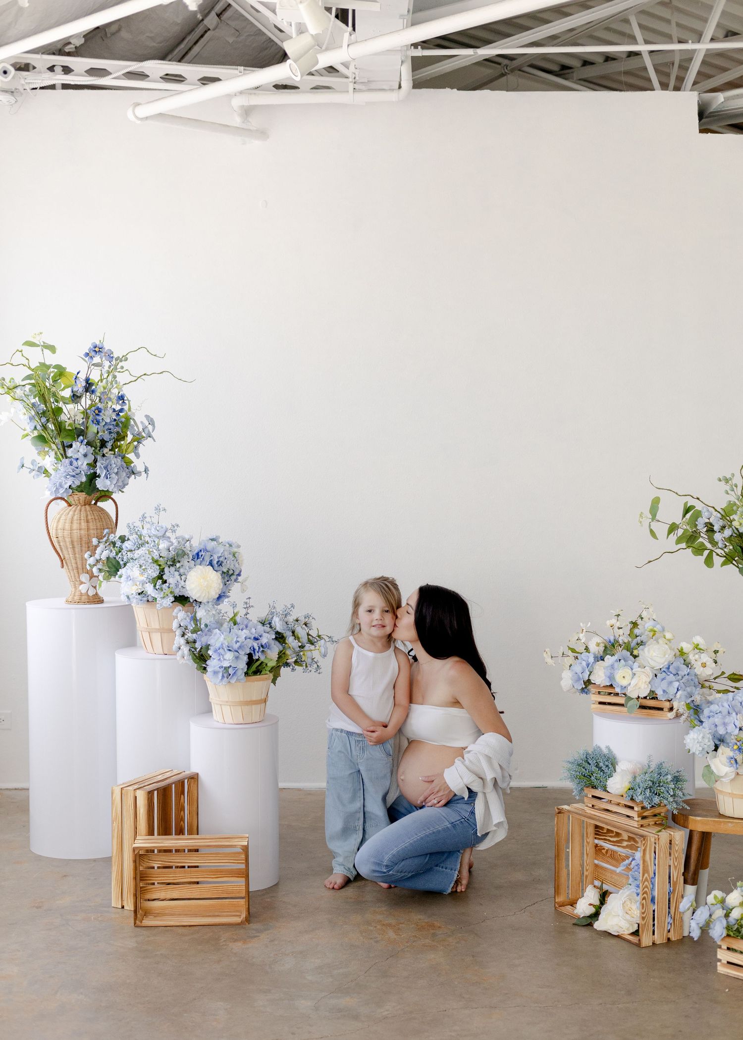 Pregnant woman crouching with young girl among blue hydrangea floral arrangements on white pedestals and wooden crates in studio.