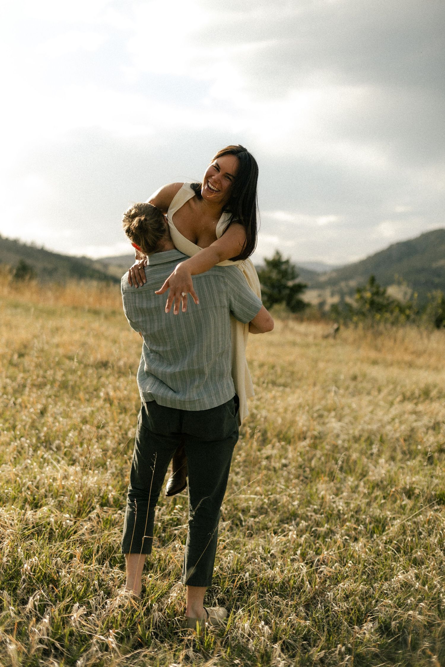 A joyful woman laughs as a man lifts her in a golden meadow with misty mountains in the background.