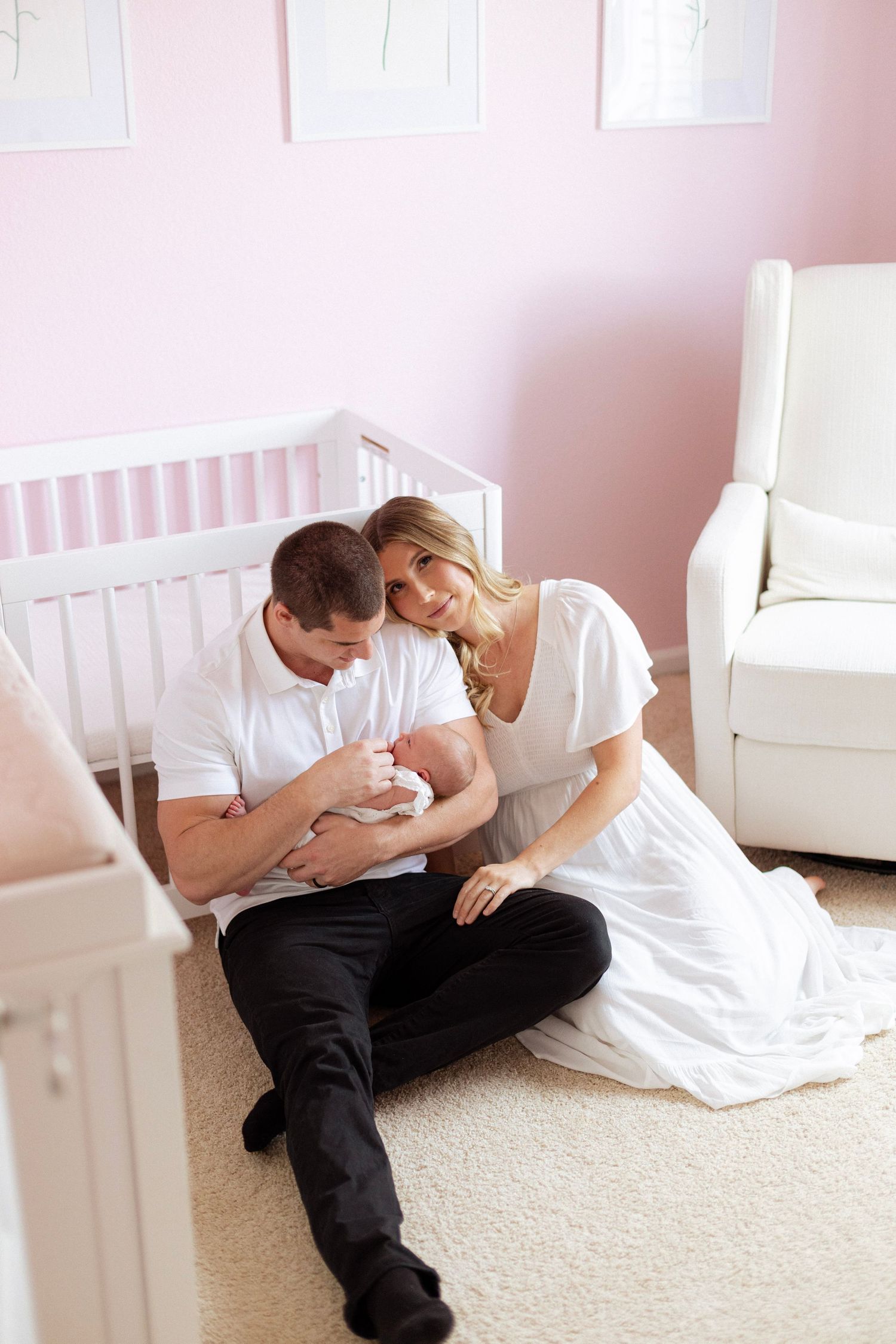 A couple sits on a nursery floor beside a white crib, tenderly holding their newborn baby in a pink room.