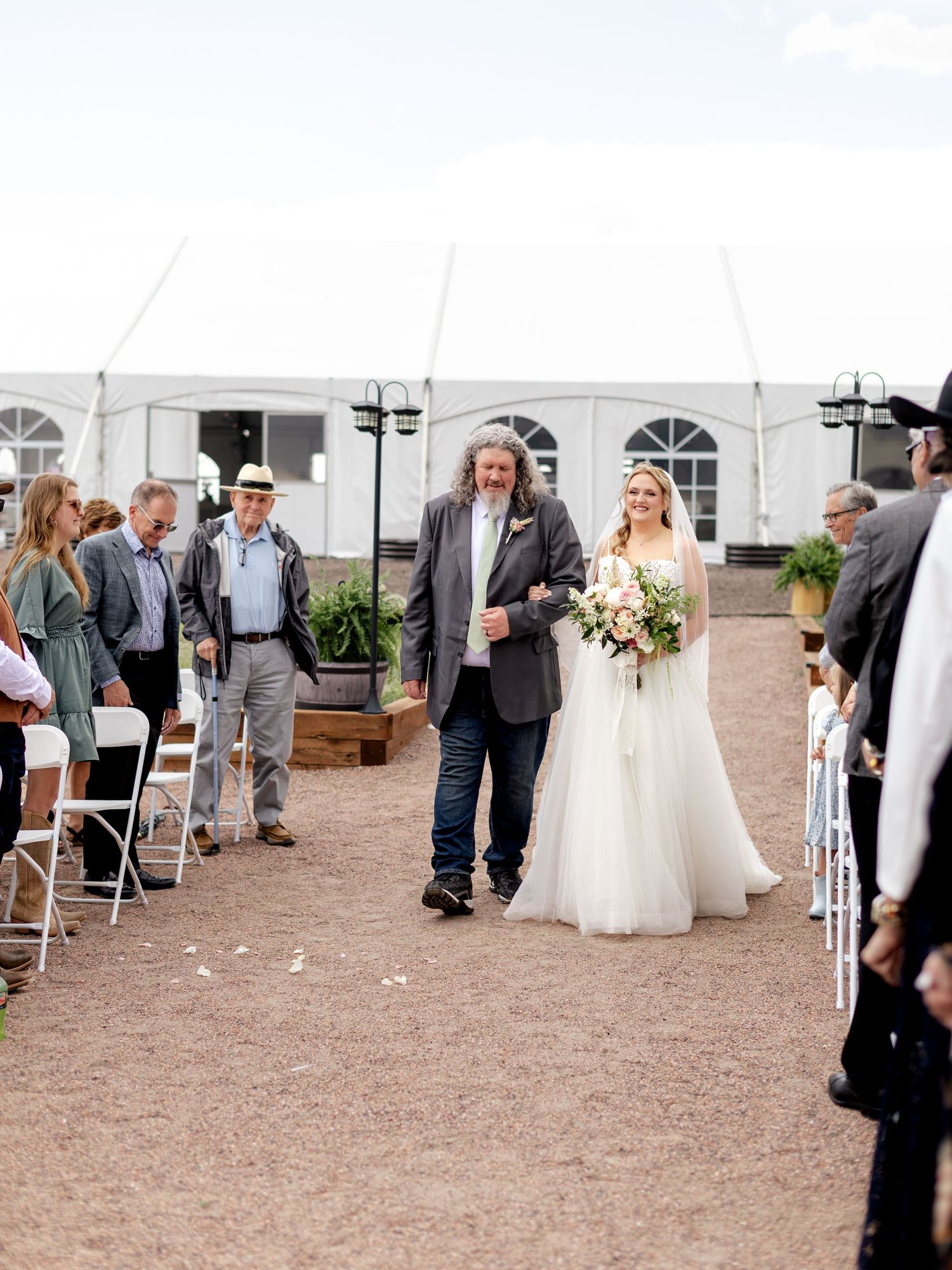 Bride walking down outdoor aisle in white gown holding bouquet escorted by her father.