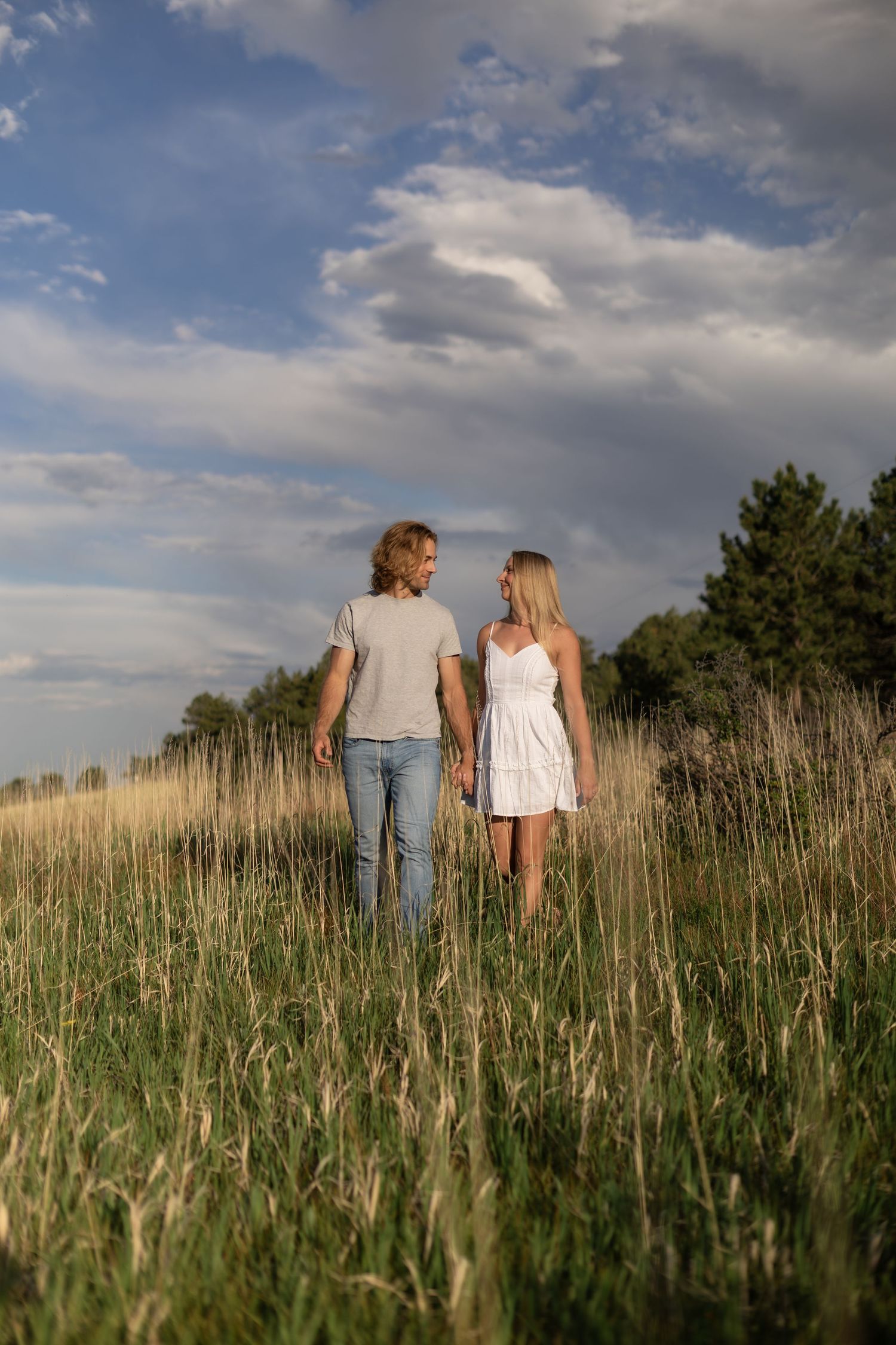 Couple holding hands walking through a golden grass field under a dramatic cloudy sky at sunset.