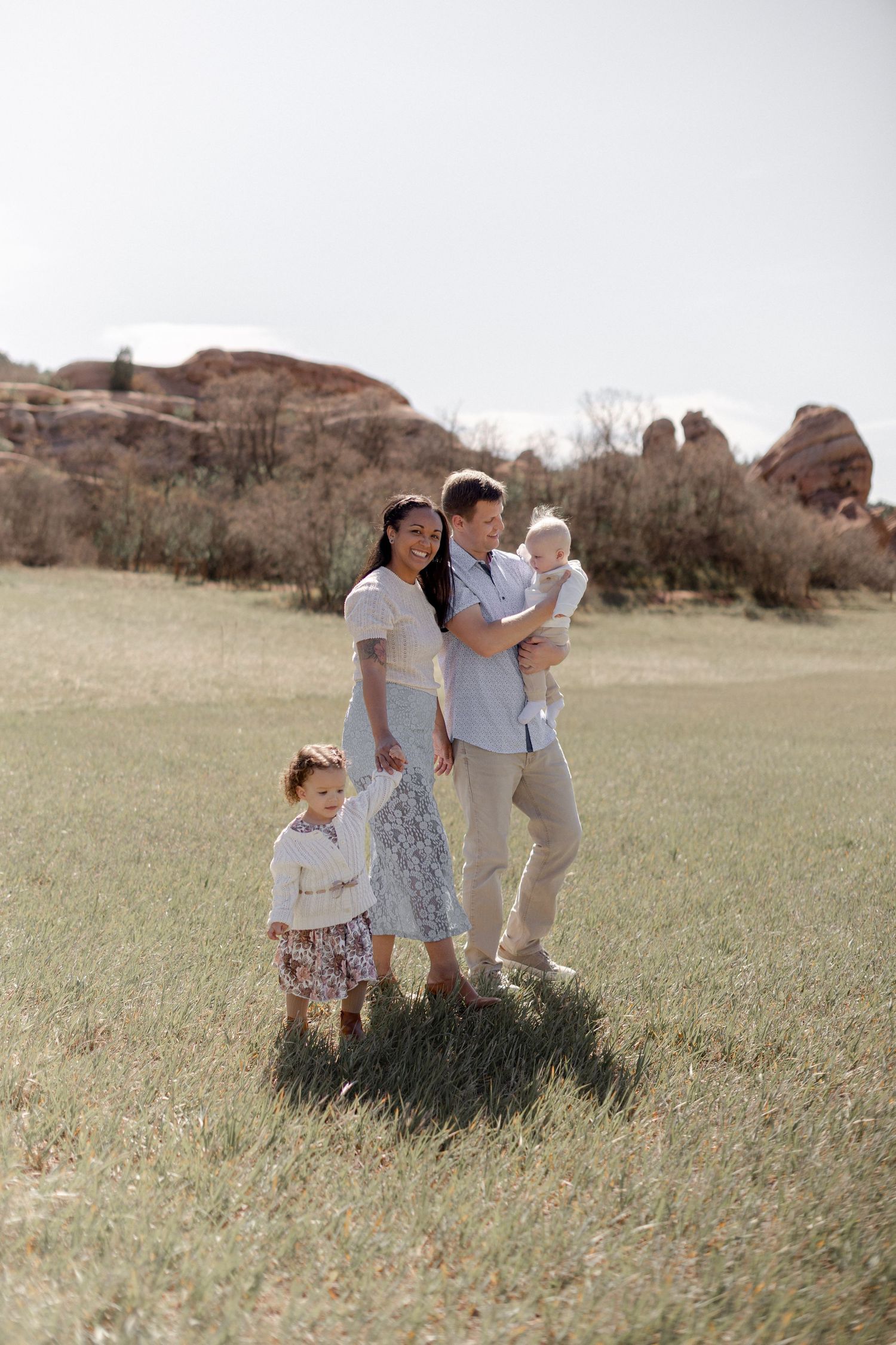 Family of four posing in a green meadow with red rock formations, parents holding children in soft neutral tones.