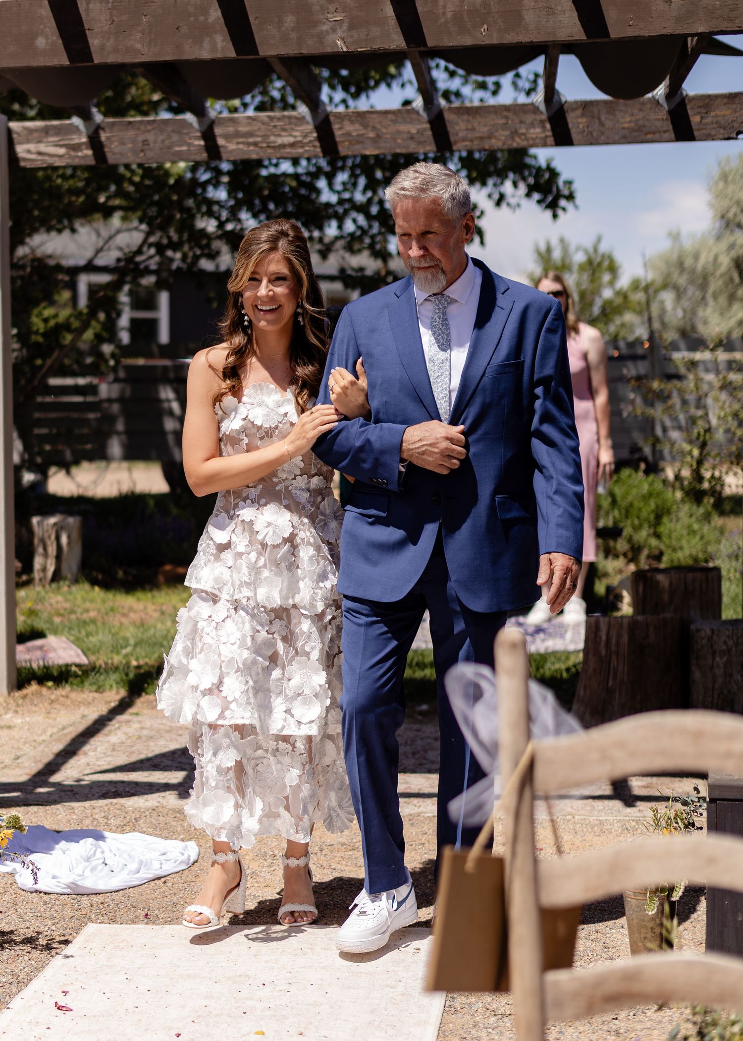 A bride in a white floral dress walks arm-in-arm with a man in a blue suit at an outdoor wedding ceremony.