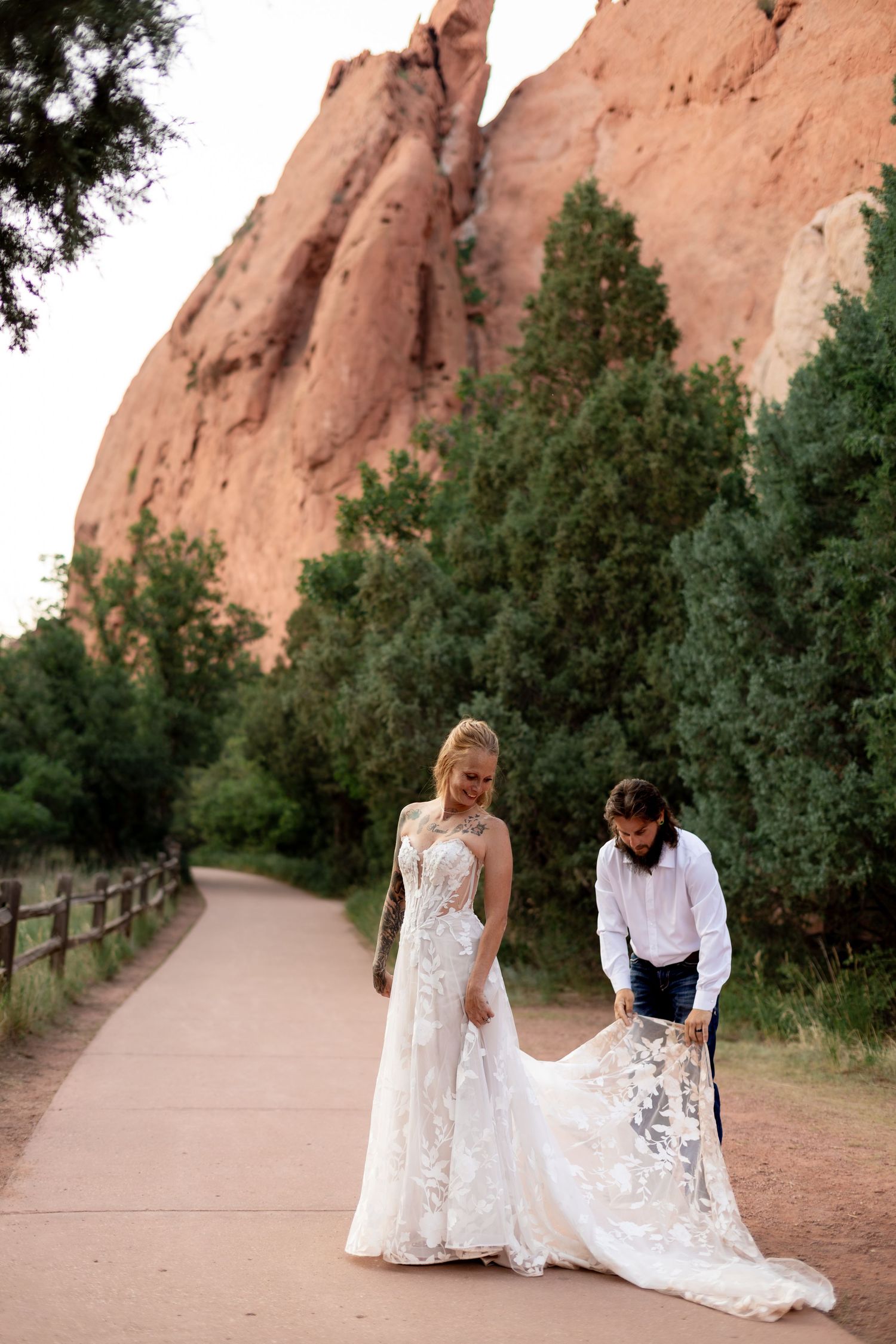 Groom adjusts bride's lace wedding dress train on a path at Garden of the Gods with towering red rock formations.