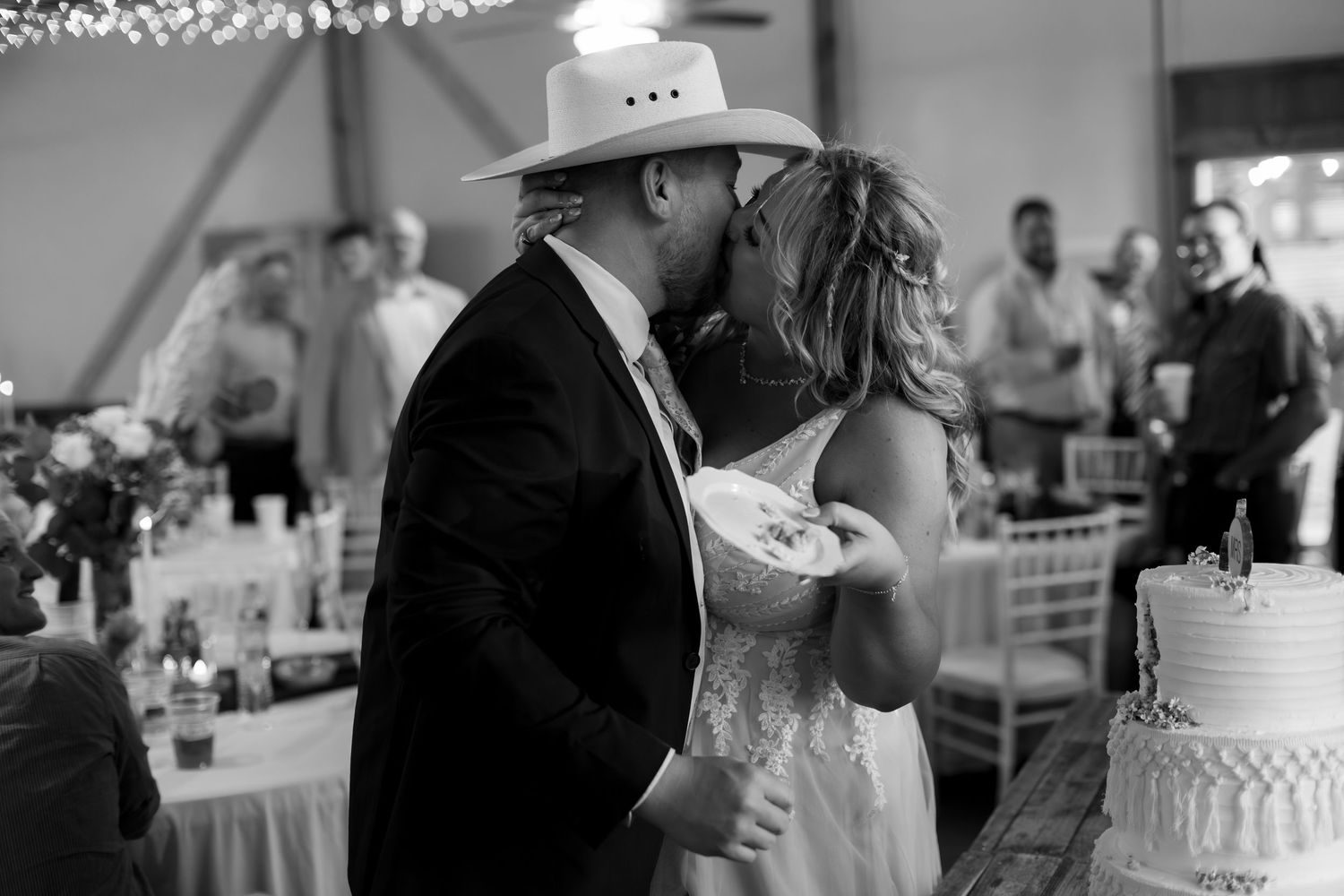 Black and white photo of bride and groom sharing a kiss during wedding cake cutting ceremony.