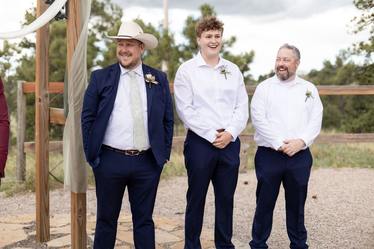 Three groomsmen in navy suits smile outdoors at a rustic wedding ceremony, one wearing a cowboy hat.