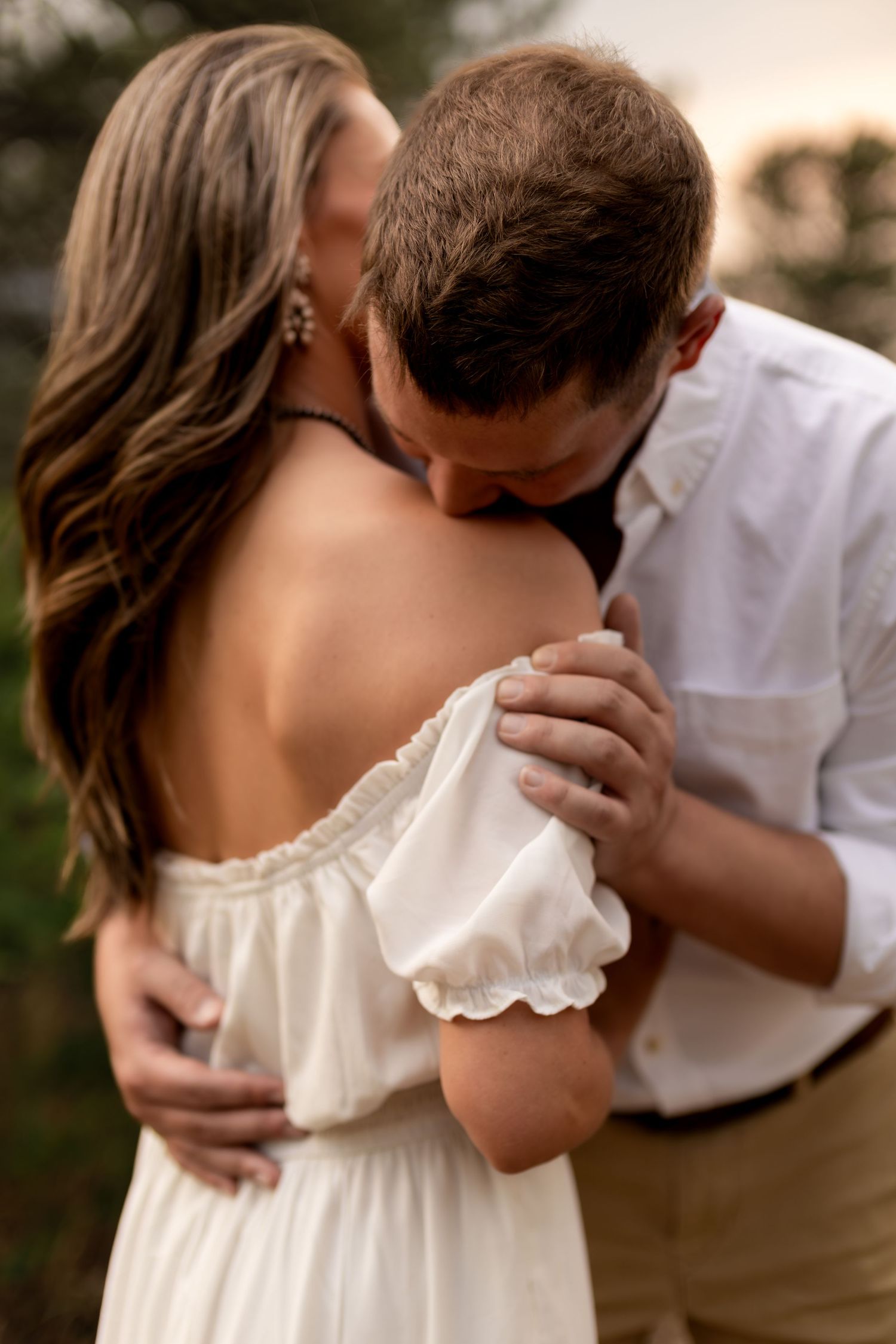 A man in a white shirt kisses a woman's shoulder as she wears an off-shoulder white dress outdoors.