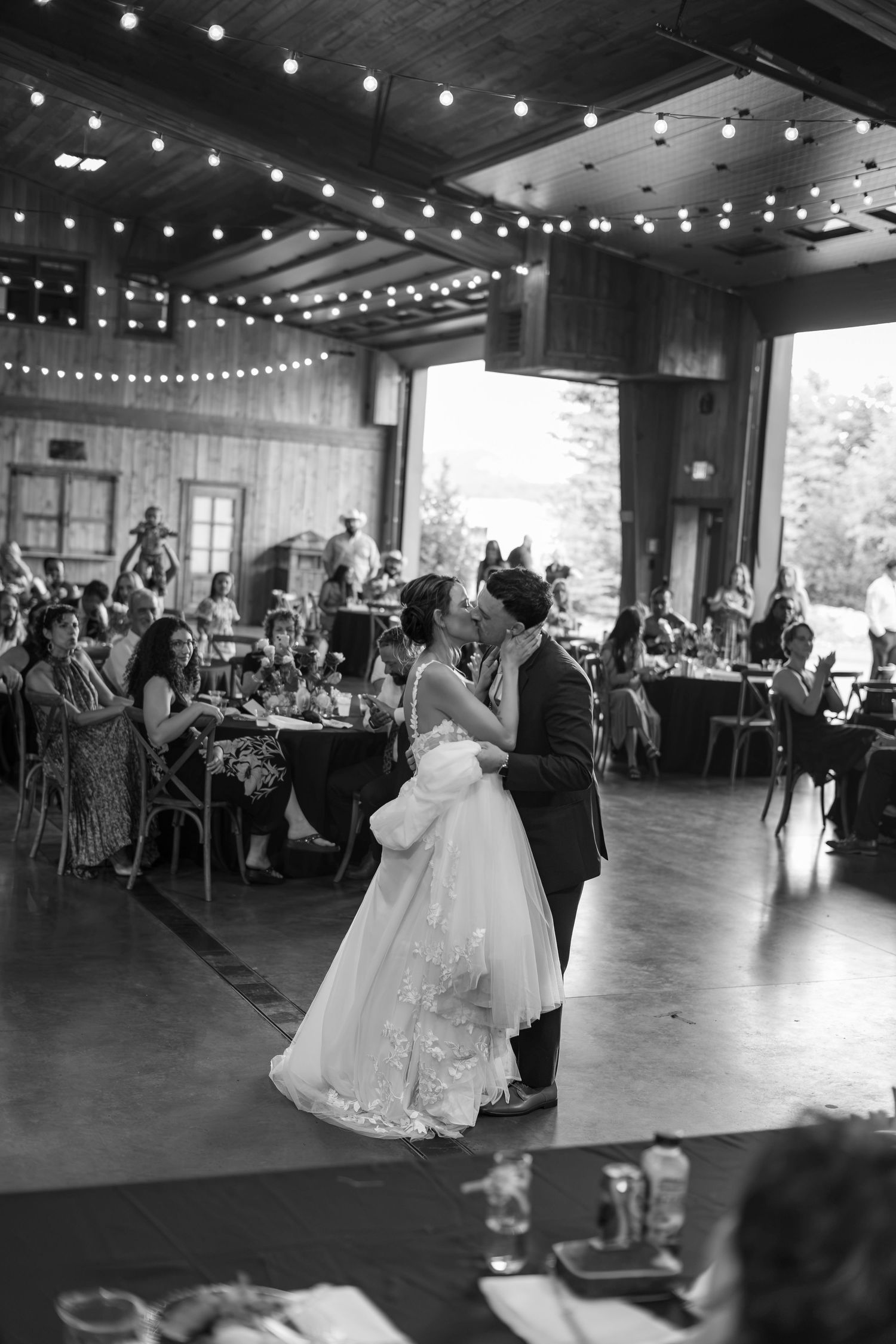Bride and groom share their first dance under string lights in a rustic barn wedding venue with guests seated around them.