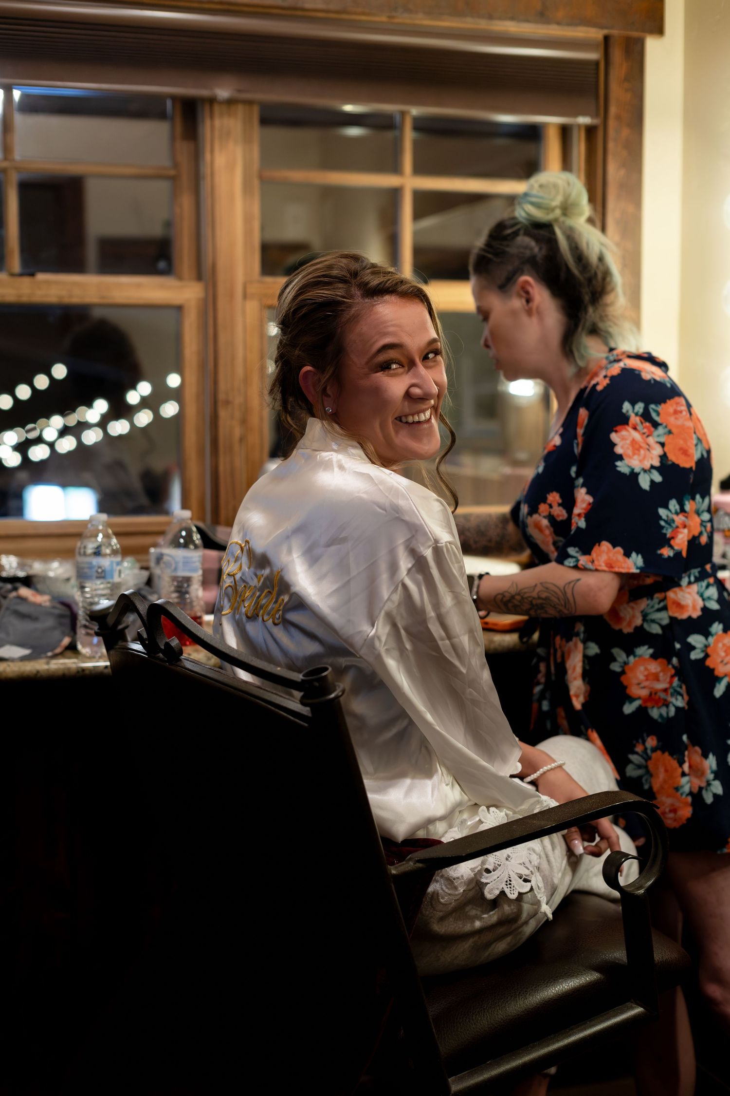 Smiling bride in white satin robe getting hair done by stylist before wedding ceremony.