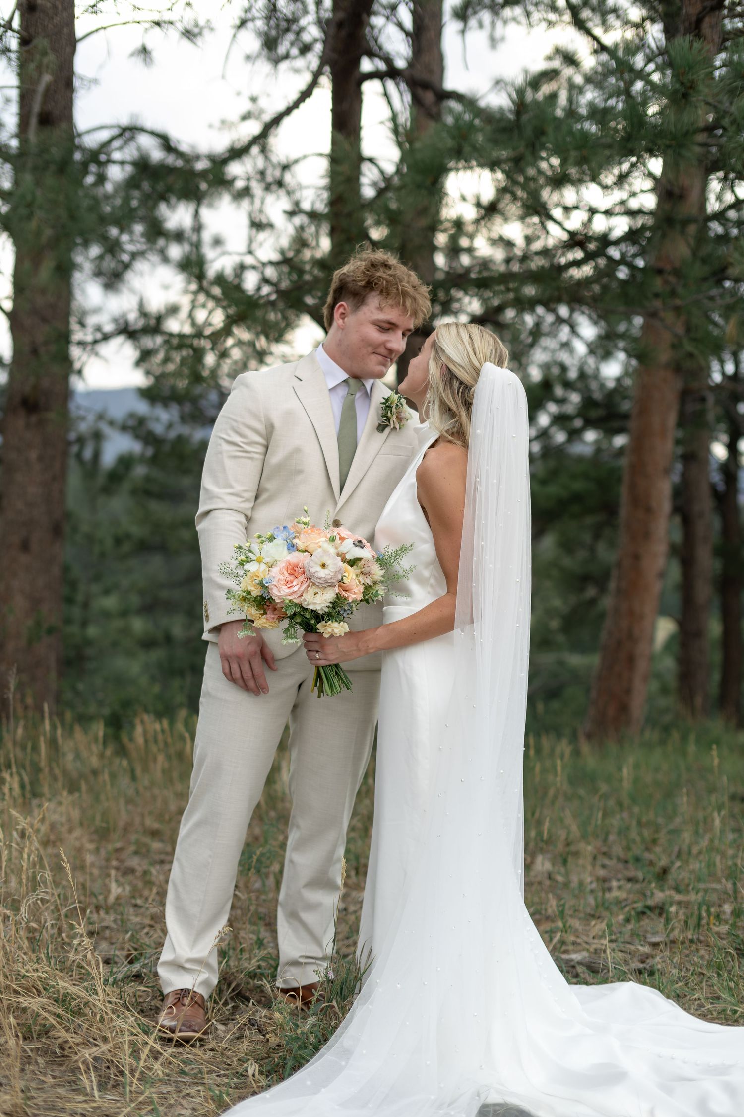 Bride and groom pose romantically among pine trees, bride holding pastel bouquet with flowing veil.