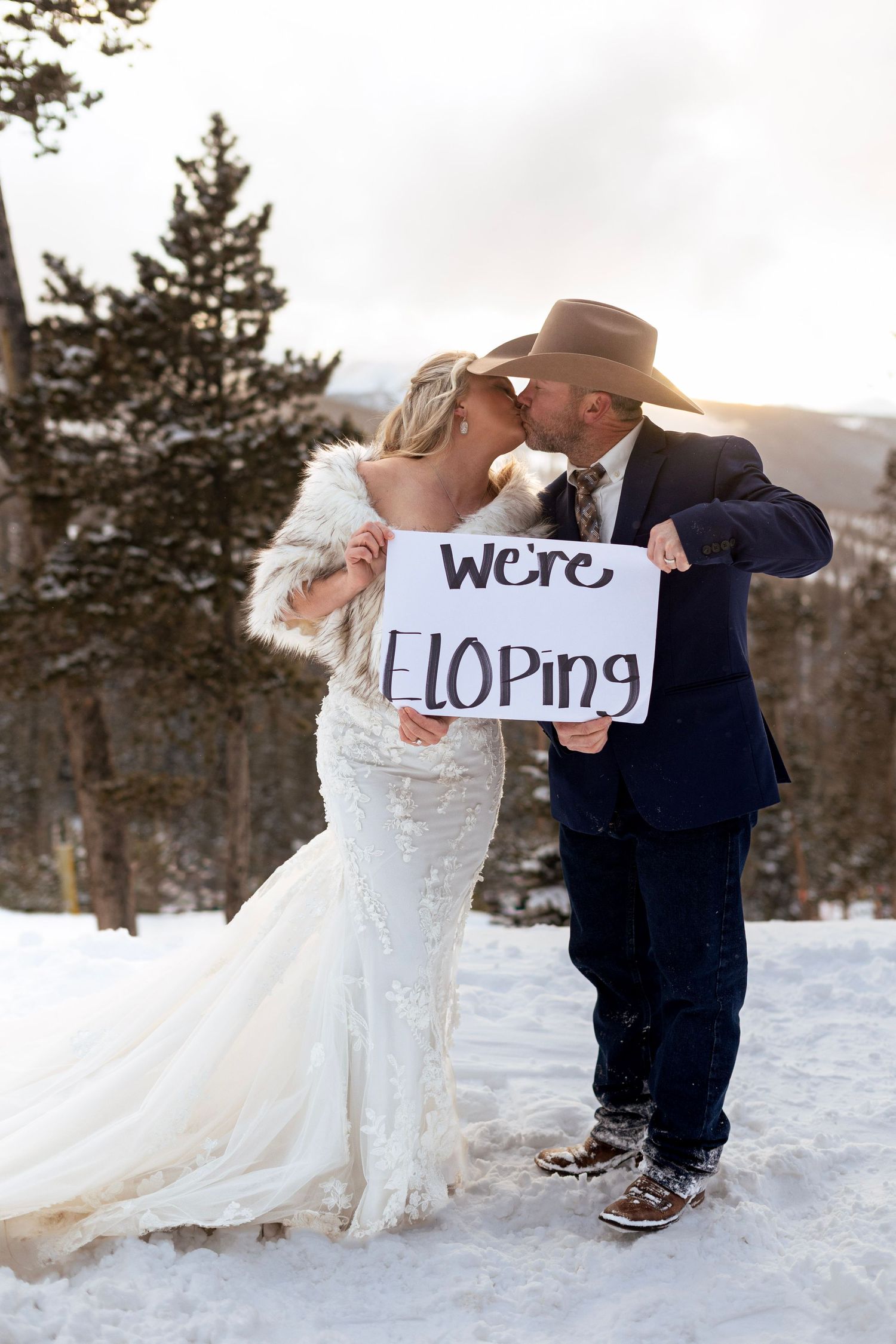 A couple in wedding attire holds up signs spelling 'ELOPING' while standing in snowy winter scenery.