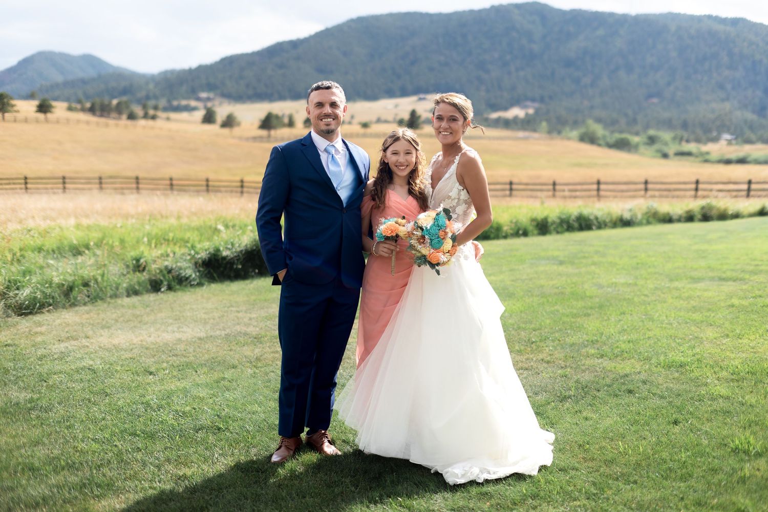 Bride and groom in navy suit pose with young flower girl in coral dress at outdoor mountain wedding.