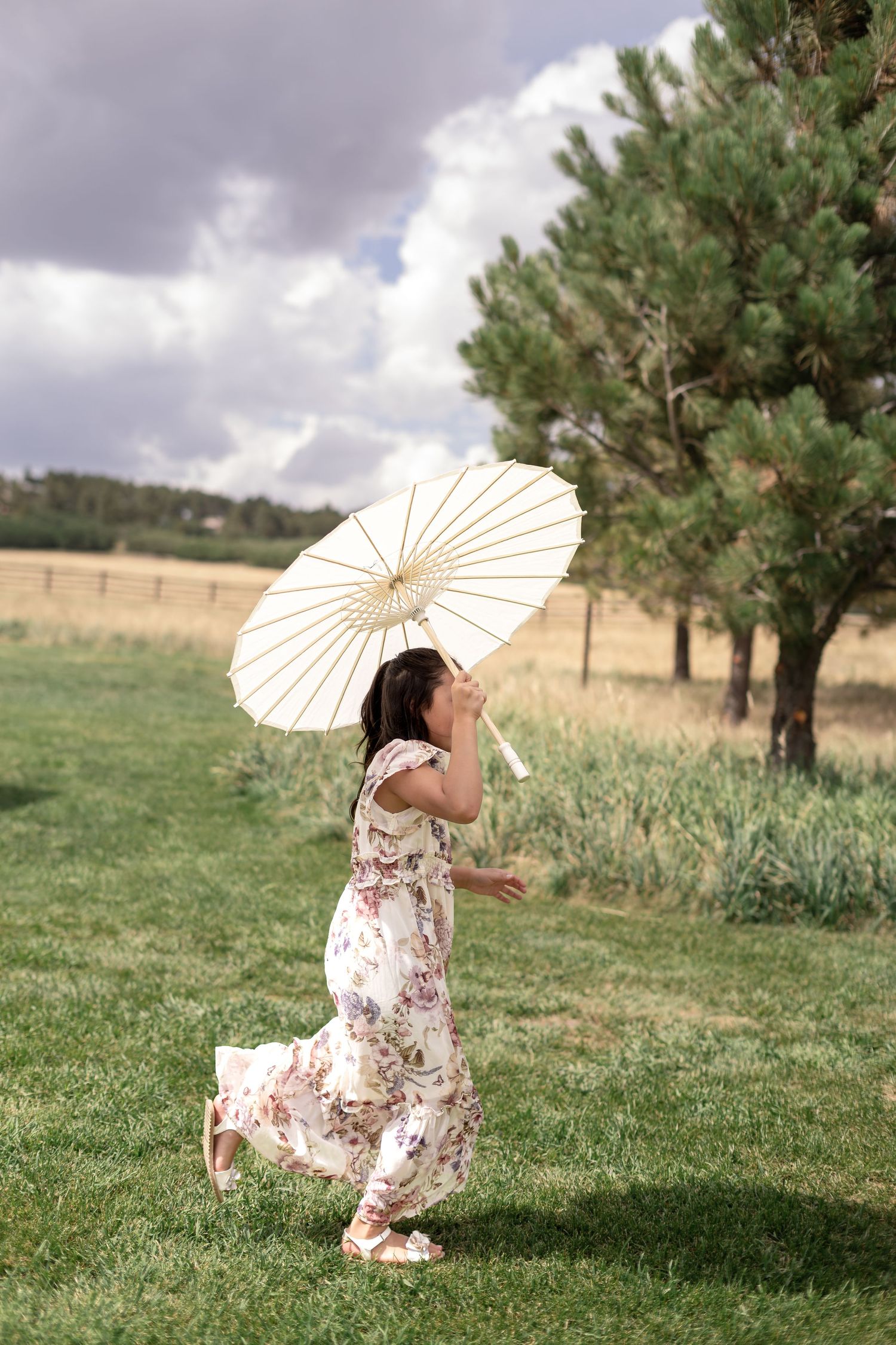 A girl in a floral dress twirls with a white parasol umbrella on green grass near pine trees.