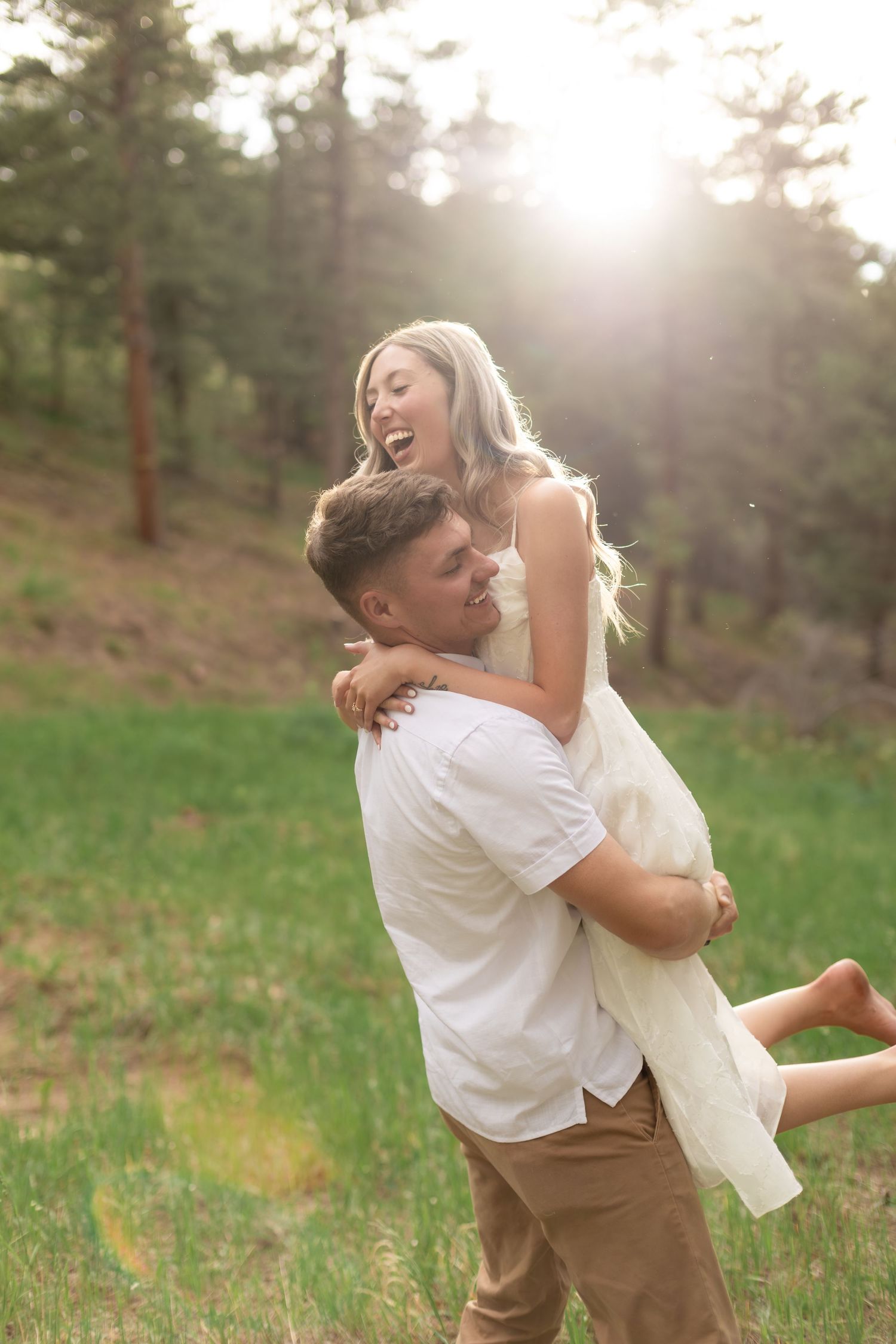 Couple laughing outdoors, man lifting woman in white dress in sunlit forest meadow.
