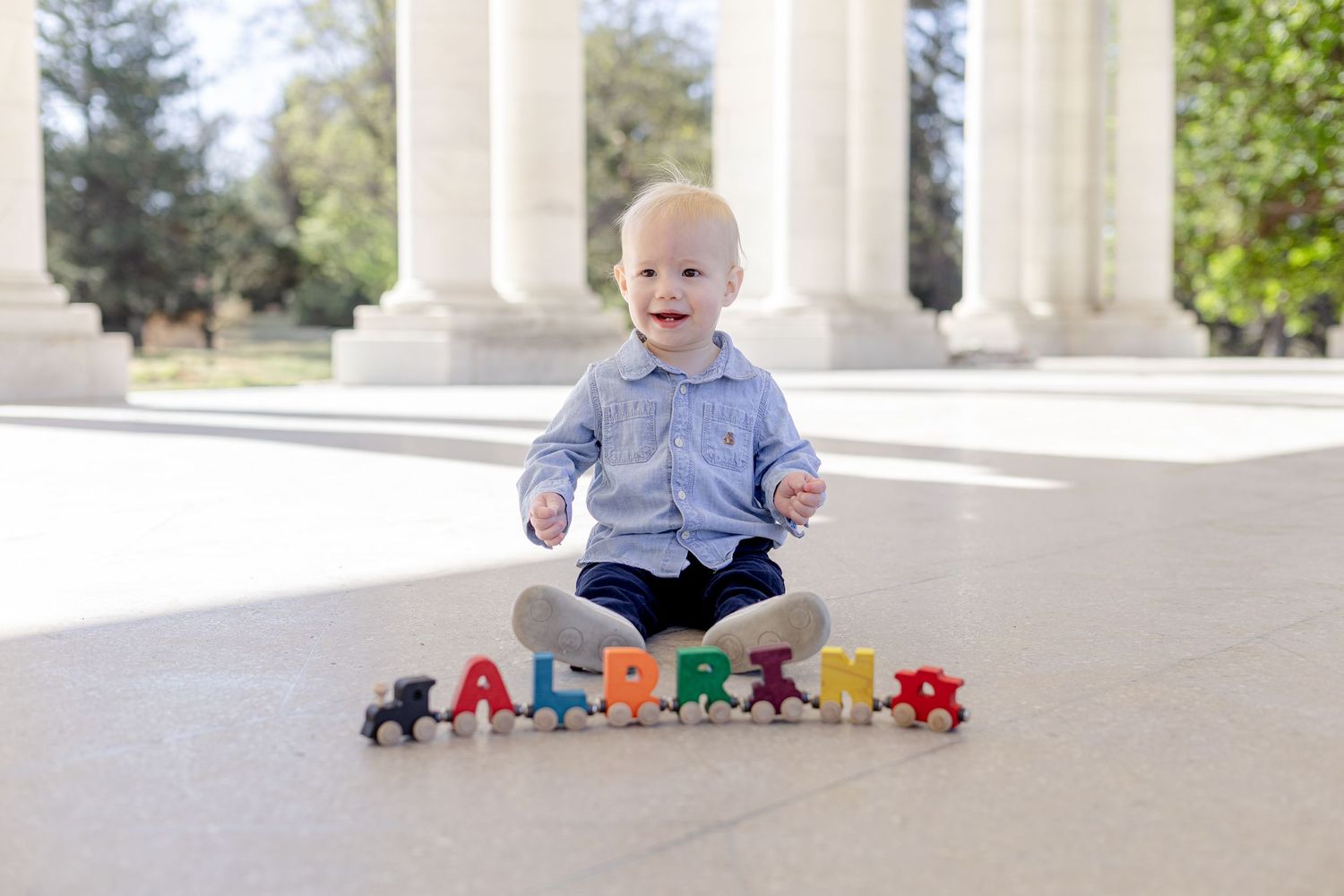 Happy toddler sitting outdoors with colorful wooden alphabet letters spelling ALPRINA on stone floor.