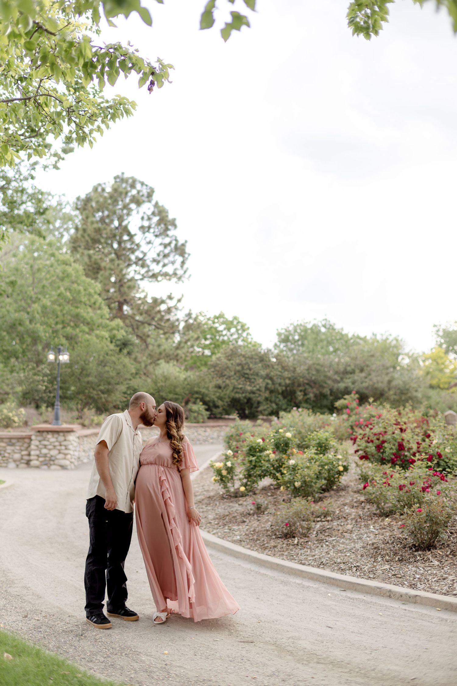 Couple sharing a kiss on a garden path, woman in flowing pink maternity dress surrounded by colorful roses.