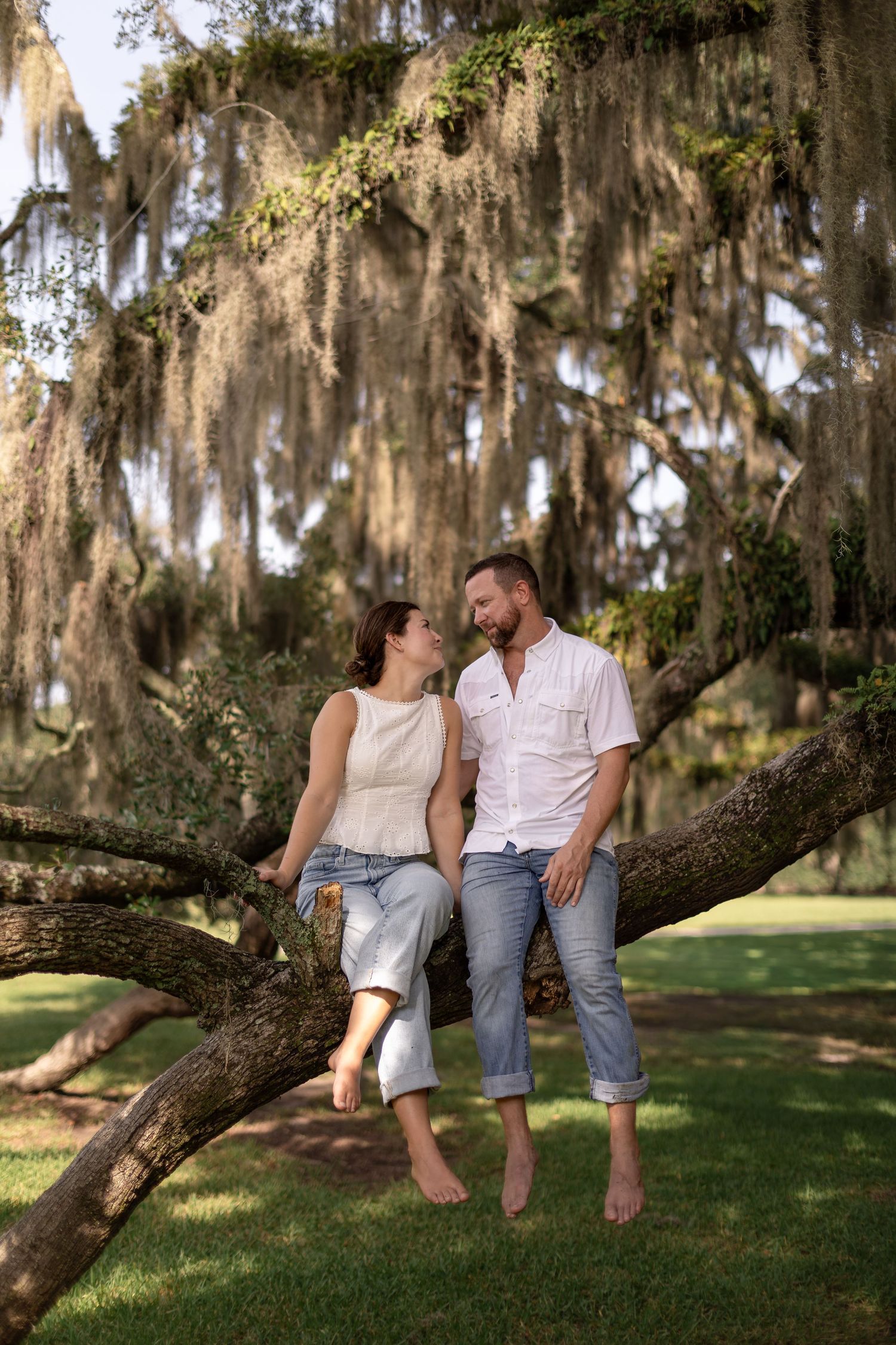 Couple sitting barefoot on a large tree branch surrounded by Spanish moss in a lush green park.