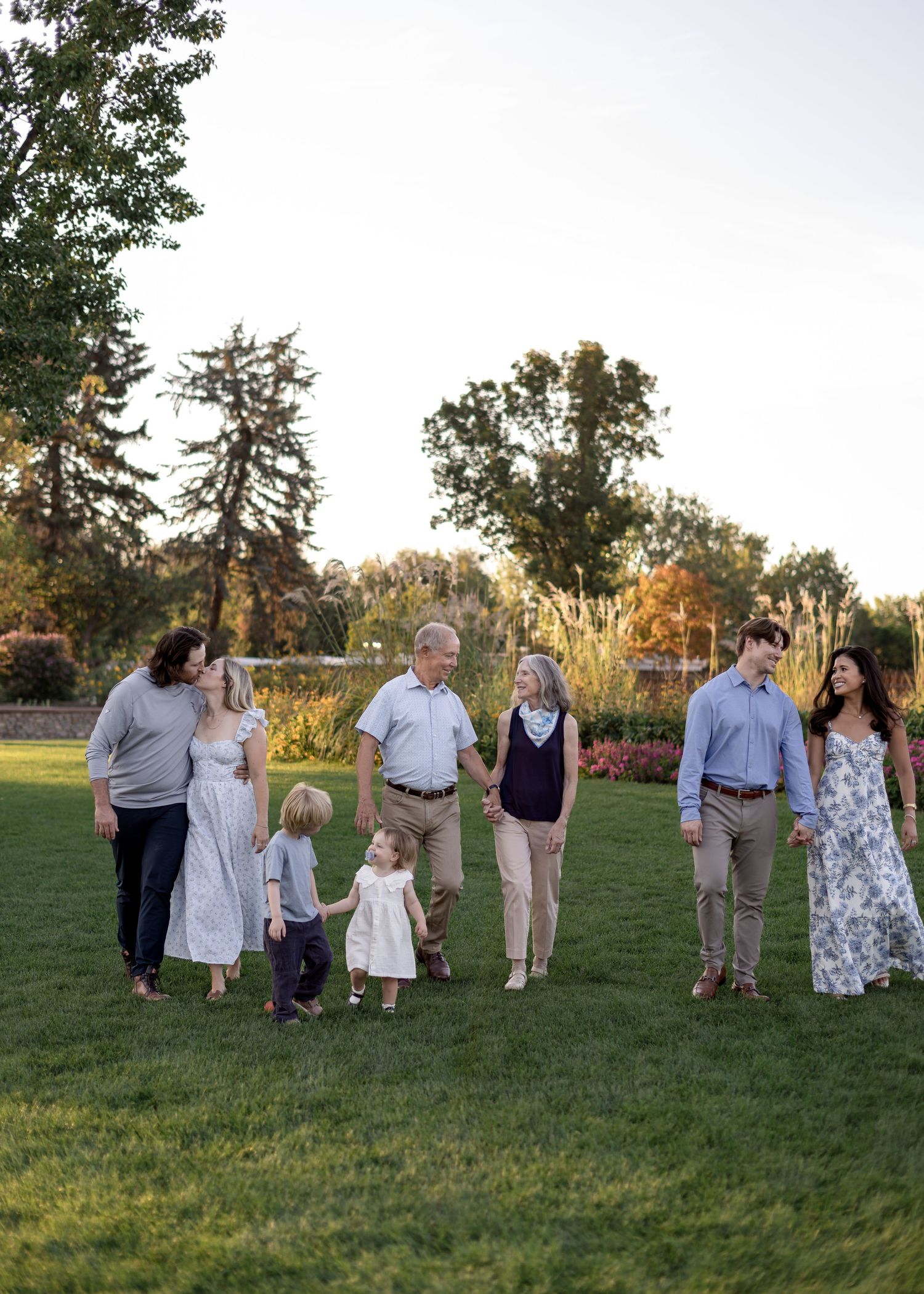 Multi-generational family walking together on lush green lawn at golden hour with trees and flowers in background.
