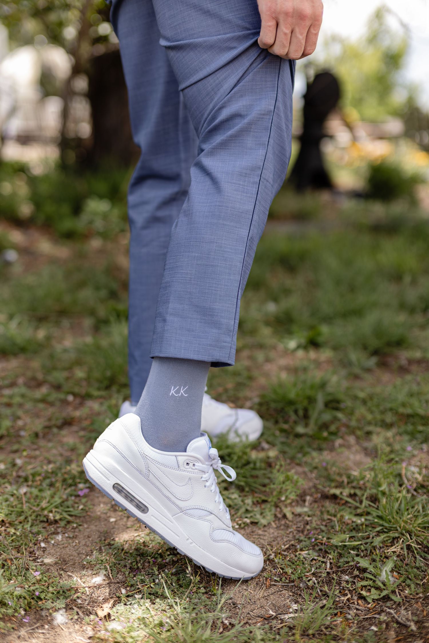 Close-up of groom's light blue dress pants with monogrammed socks and white Nike Air Max sneakers outdoors.