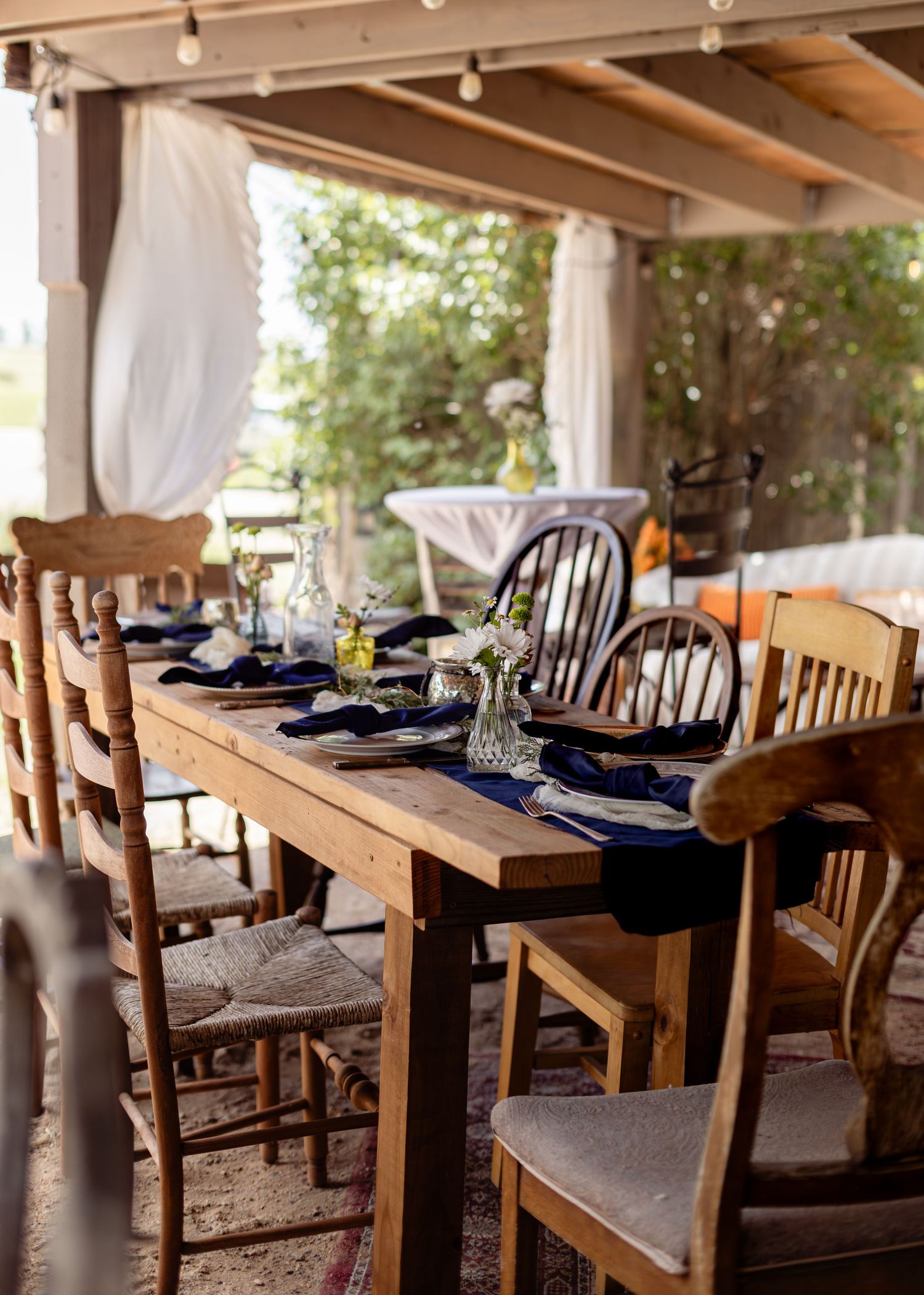 Rustic outdoor dining table set with navy napkins and wooden chairs under a pergola with white curtains.