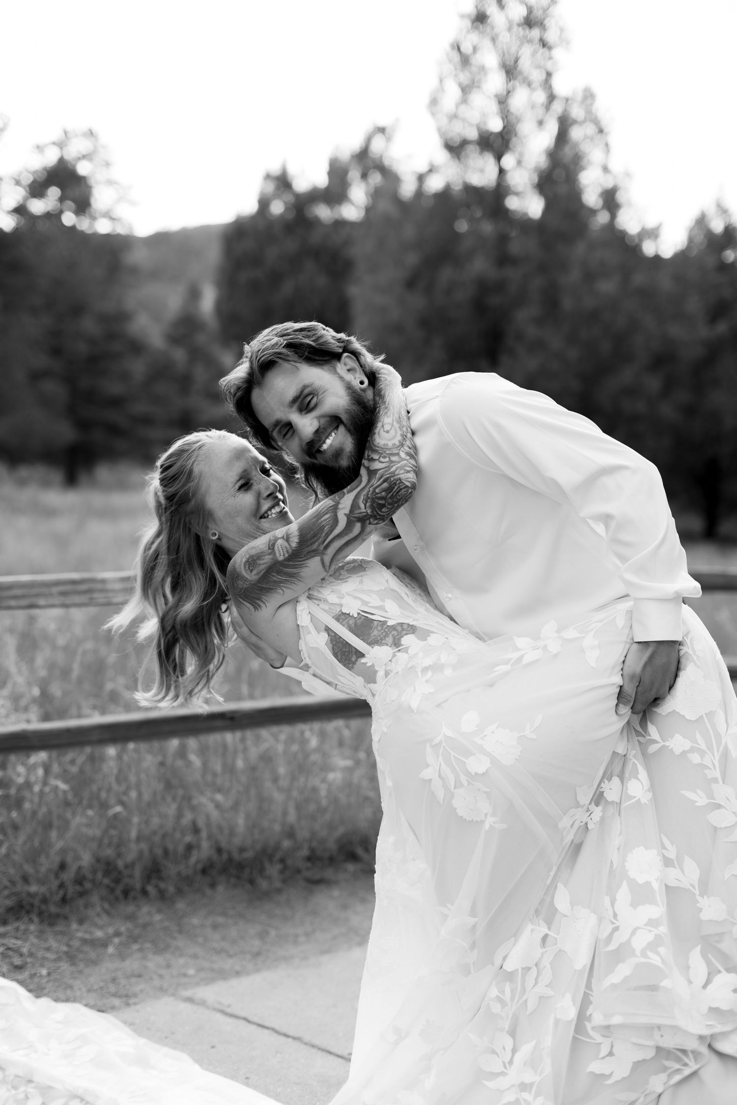 Black and white photo of joyful bride and tattooed groom sharing playful embrace at outdoor wedding.