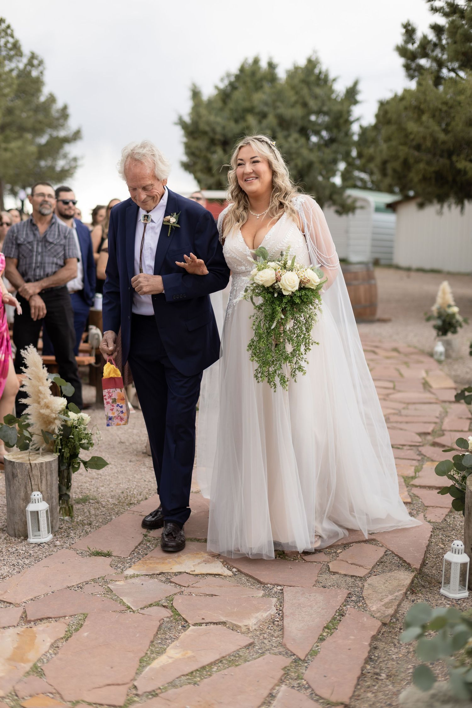 Smiling bride in a white tulle gown holding a cascading bouquet walks down the aisle with an older gentleman.
