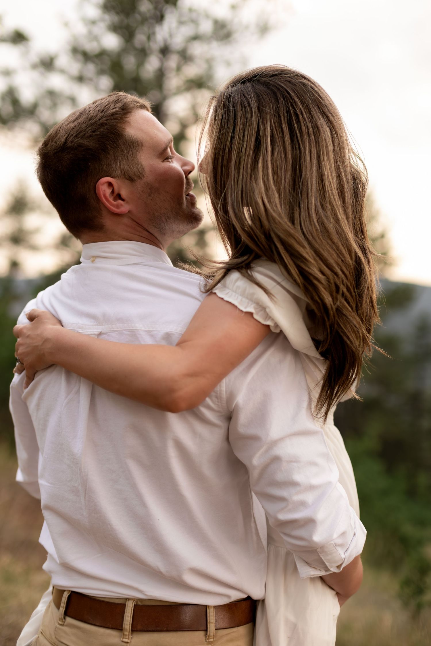 Couple embracing and kissing outdoors during a romantic engagement photo session with pine trees in the background.