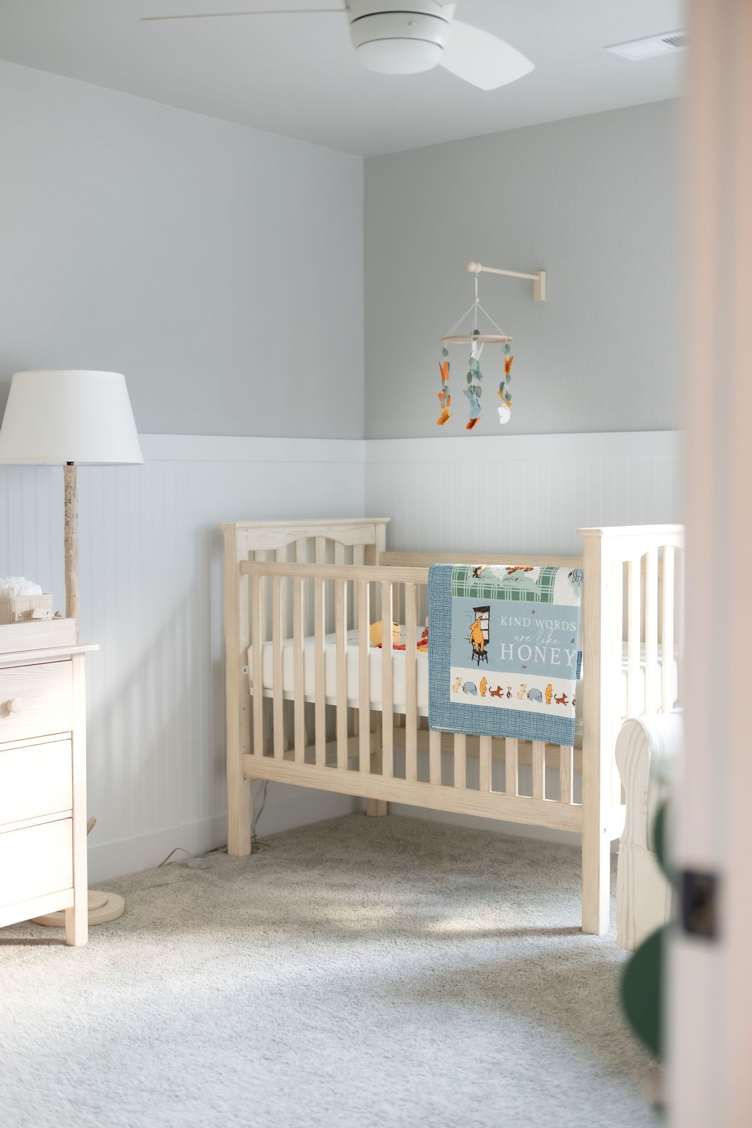Serene baby nursery with white crib, mobile, lamp, dresser, and blue Winnie the Pooh blanket on gray carpet.