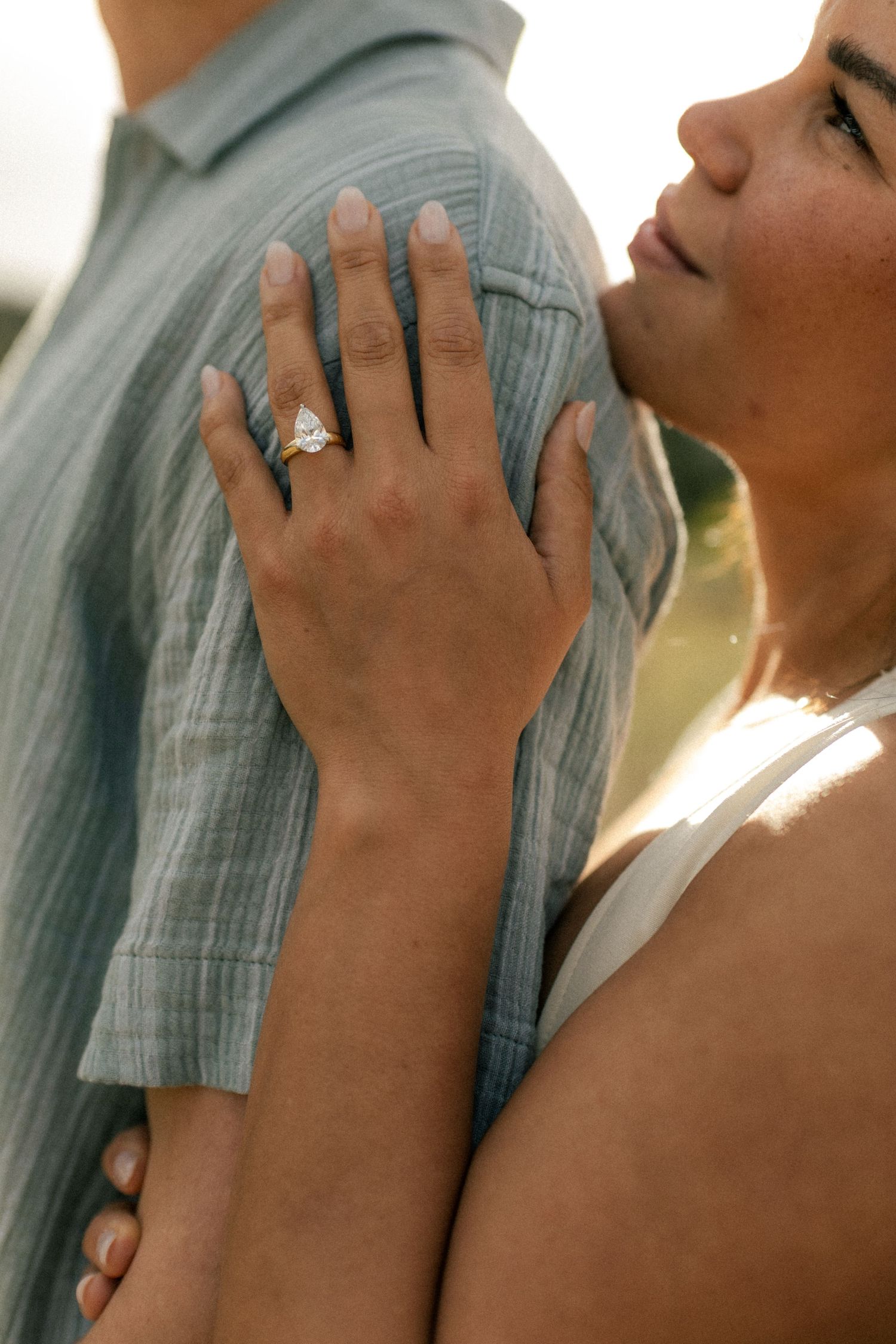 Close-up of womans hand with pear-shaped diamond engagement ring resting on mans blue linen shirt.