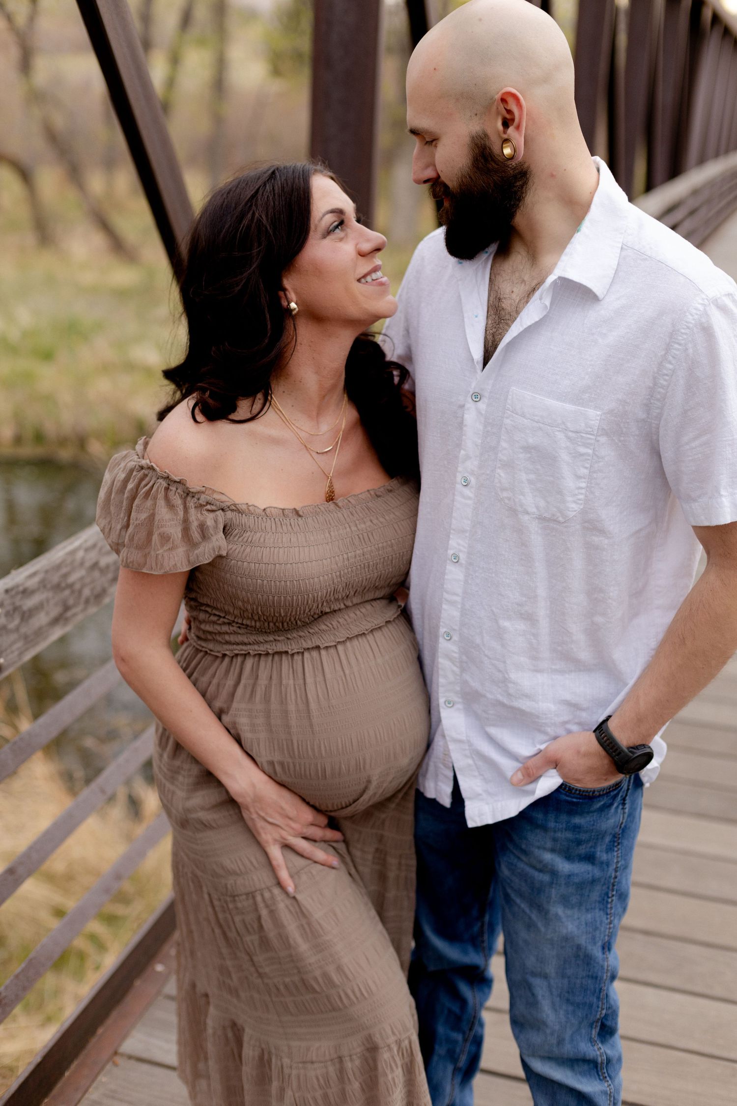 Pregnant woman in brown off-shoulder dress with bald bearded man in white shirt on a bridge, maternity photo shoot.