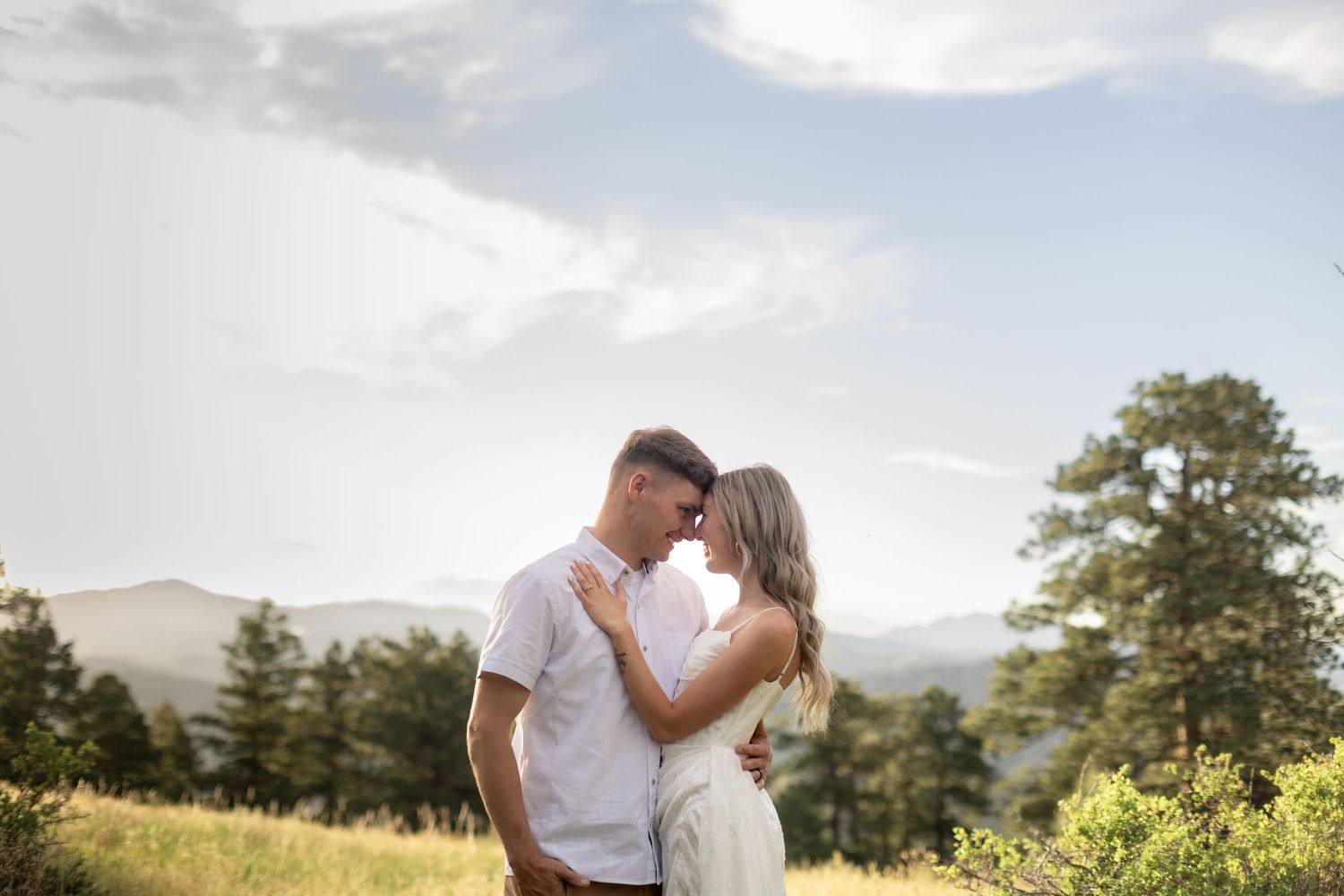 Couple embracing on a scenic mountain meadow with pine trees and cloudy sky in the background.